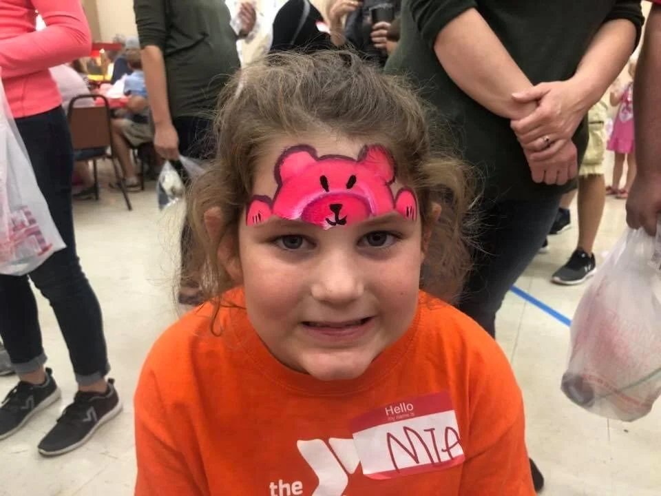 A young girl with curly hair, wearing an orange shirt and a name tag that reads 'Nia', has a pink face paint design of a bear on her forehead, and is smiling at the camera in a crowded indoor setting.