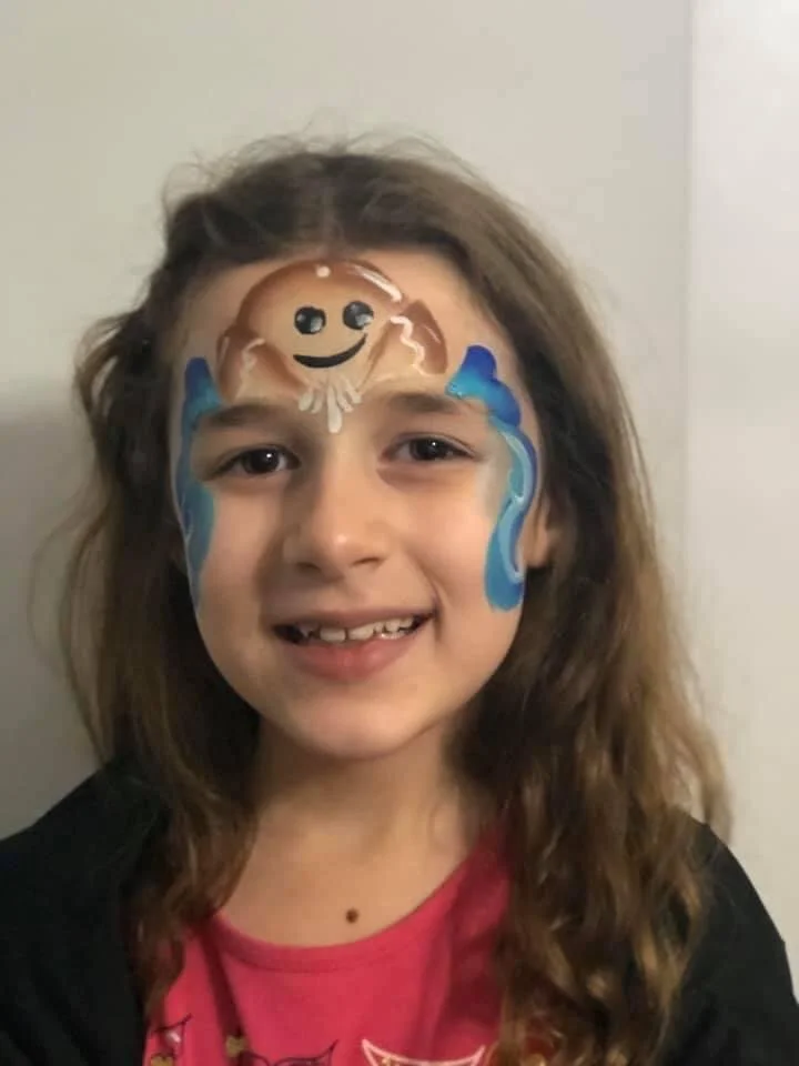 Child with a painted face featuring a smiling brown octopus with blue water splashes on cheeks.