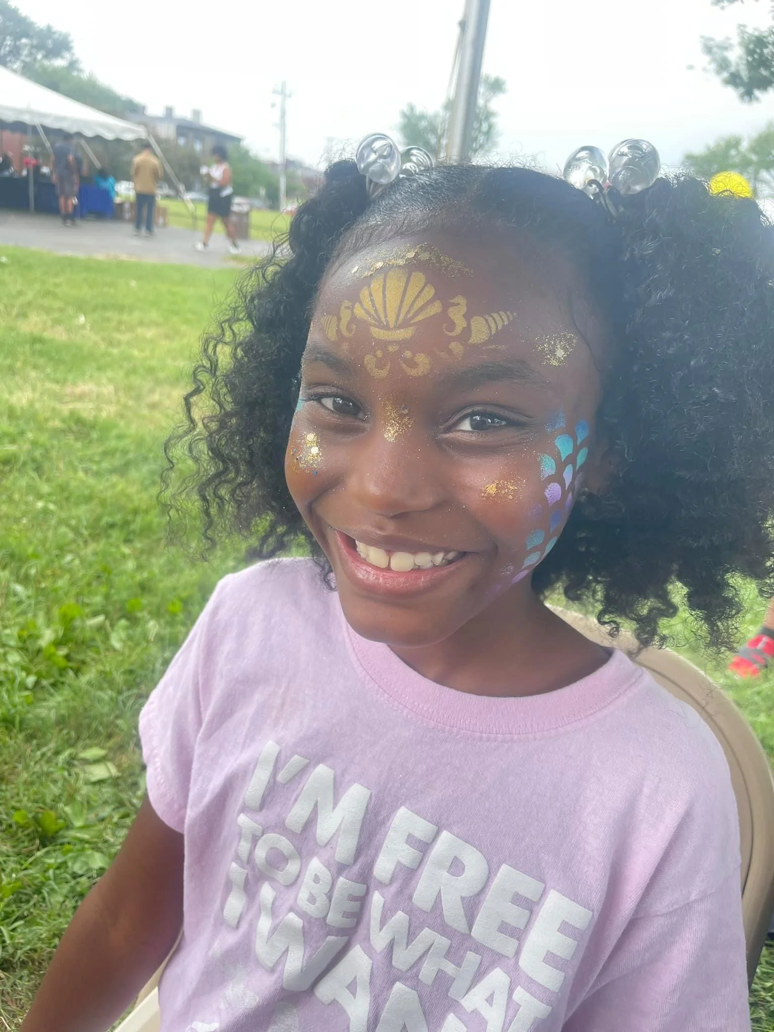 A young girl with curly hair smiles at the camera, her face decorated with colorful face paint and gold accents, outdoors at an event or festival.