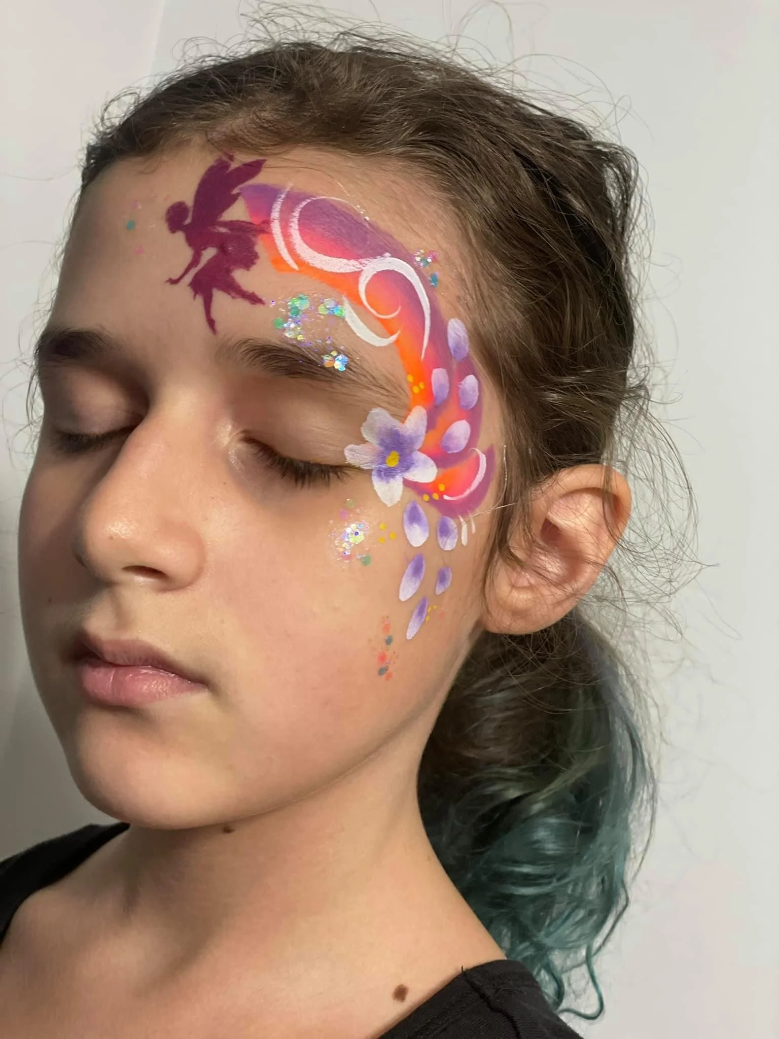 A young girl with closed eyes features a colorful face painting of a fairy with purple, pink, and orange hues, including flowers, swirling patterns, and glitter on her right temple.