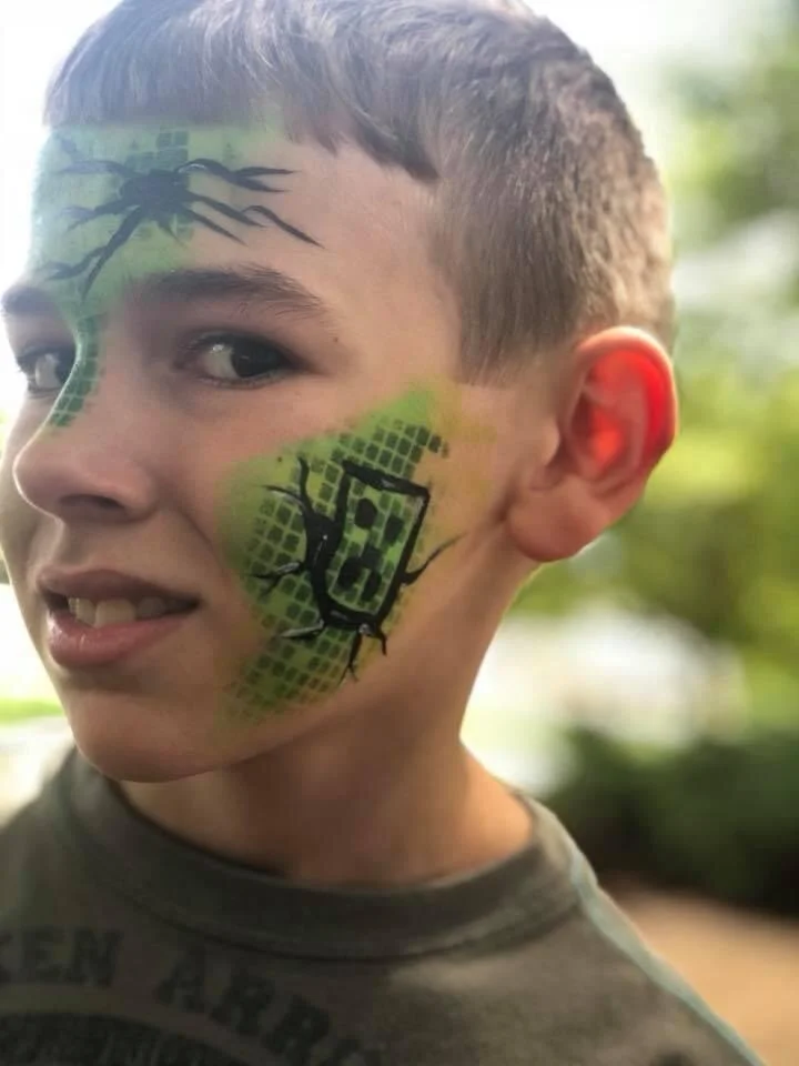 Close-up of a young boy with face painted in green and black, featuring spider and web designs. He has short hair, a slight smile, and appears to be outdoors with blurred greenery in the background.