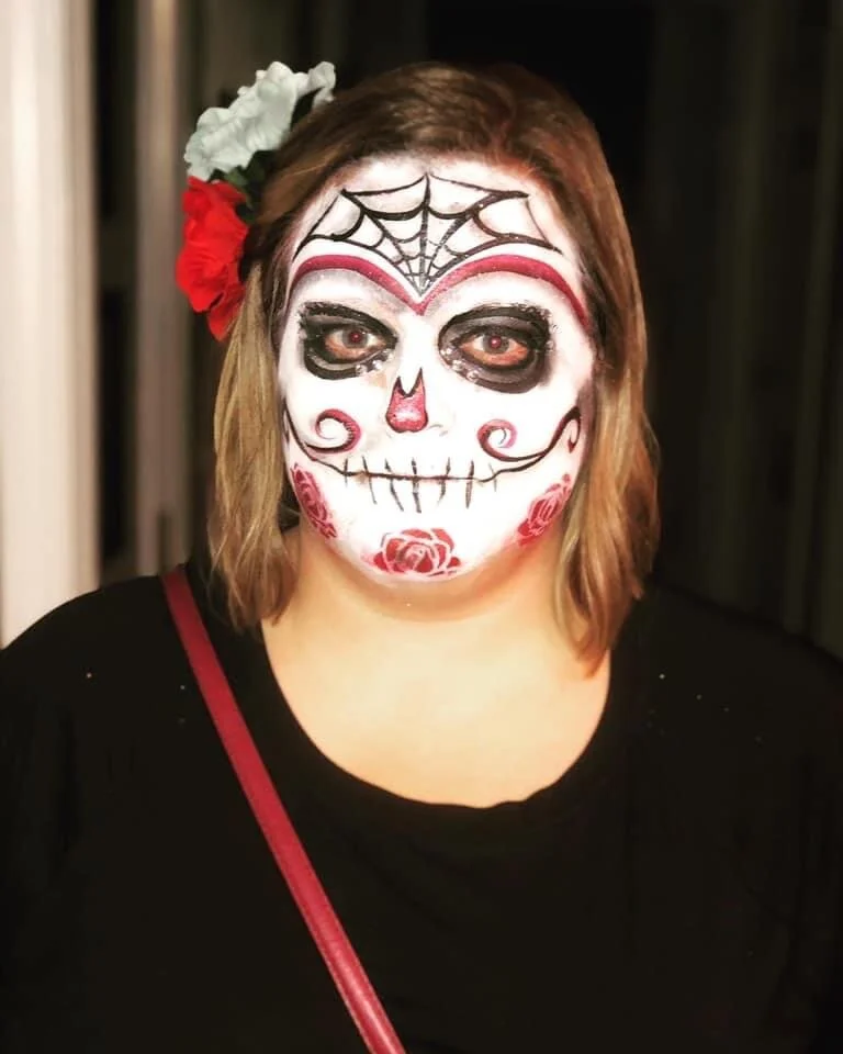 Woman with Day of the Dead sugar skull face paint, decorated with roses and spider web designs, wearing black clothing and a flower hair accessory.