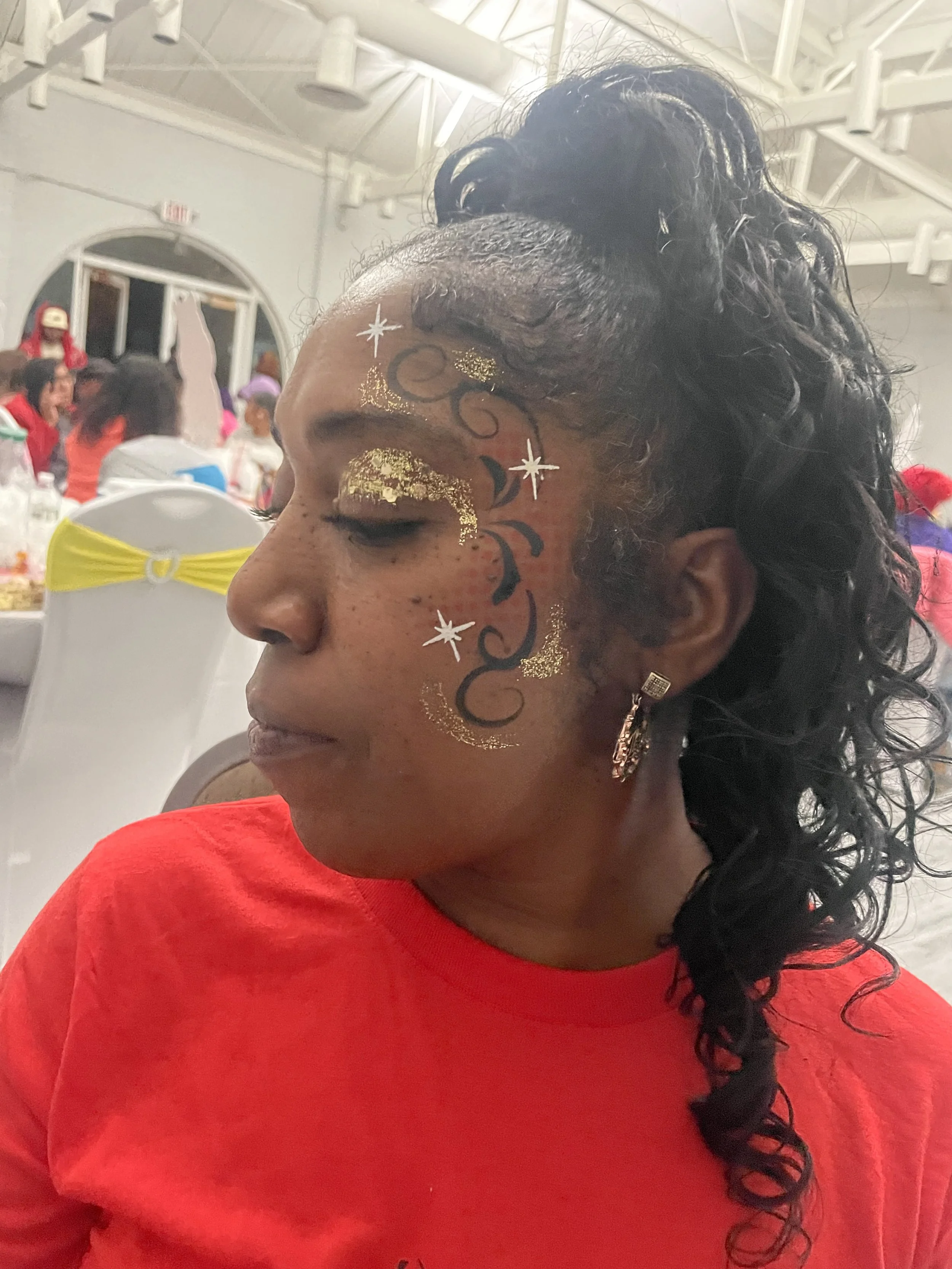 A woman with dark curly hair styled in a high ponytail, wearing a red shirt and silver earrings, with decorative face paint and gold glitter around her eye in a festive indoor setting.