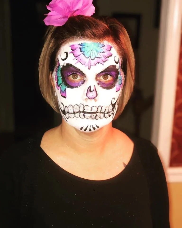 A woman with face paint resembling a colorful sugar skull, wearing a pink flower hair accessory, in a black top.