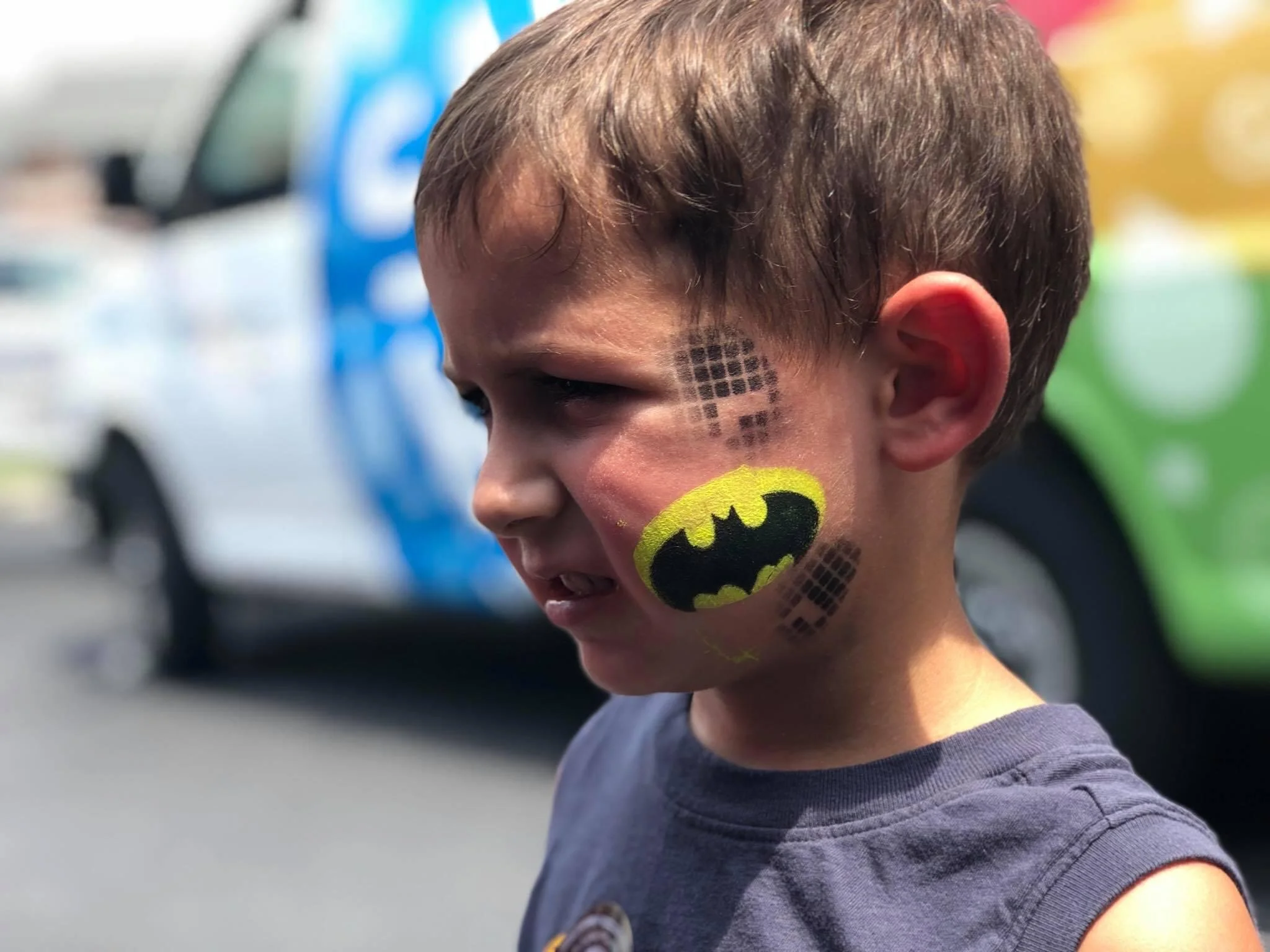 Side profile of a young boy with face paint of Batman logo on his cheek, with a black dotted heart pattern above it, standing outdoors with a blurred police car in the background.