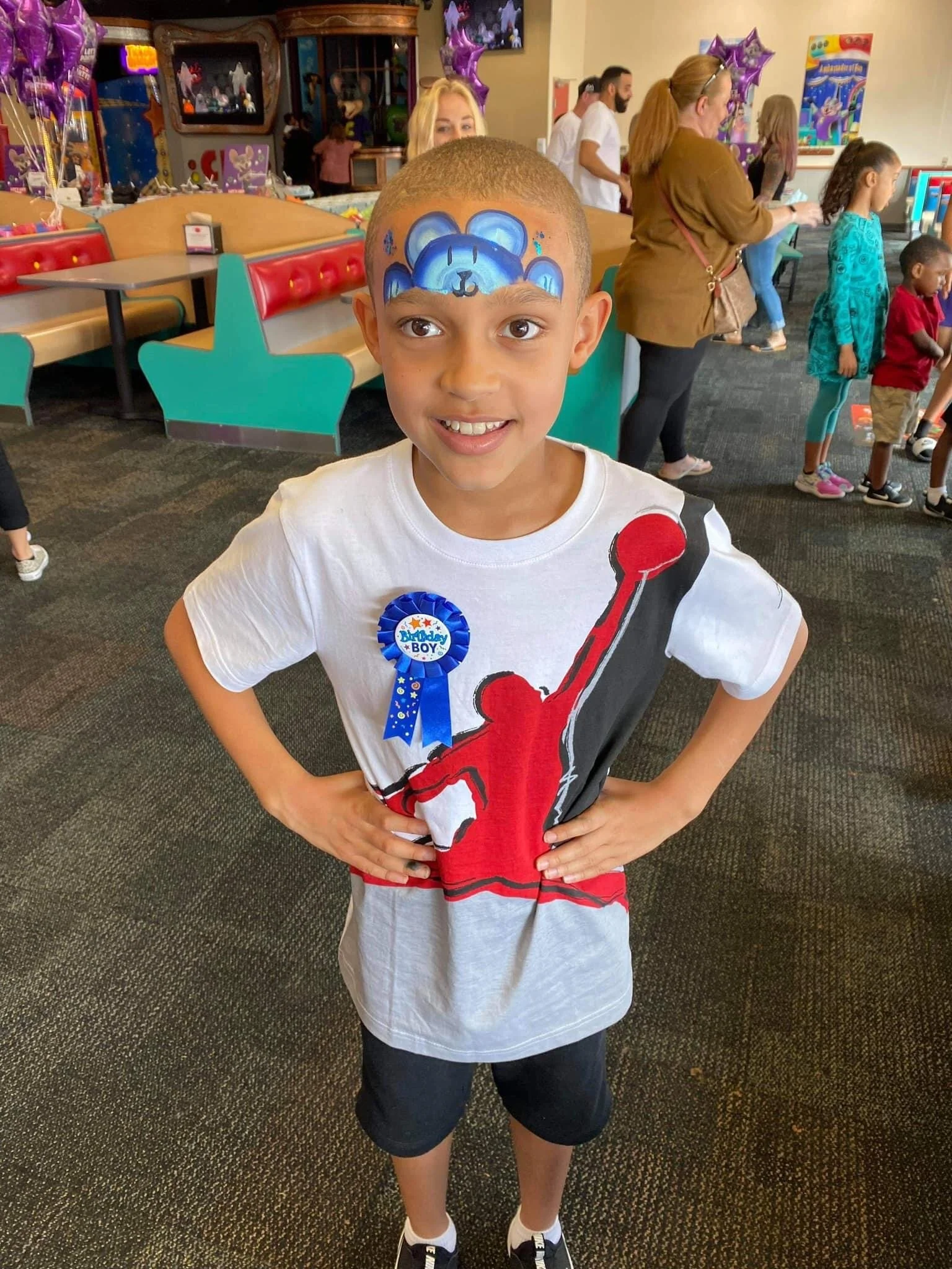 A young boy with a Buzz Lightyear face painting, standing with hands on hips inside a colorful arcade or entertainment center, wearing a white t-shirt with a red and black basketball graphic, a blue badge, black shorts, and sneakers.