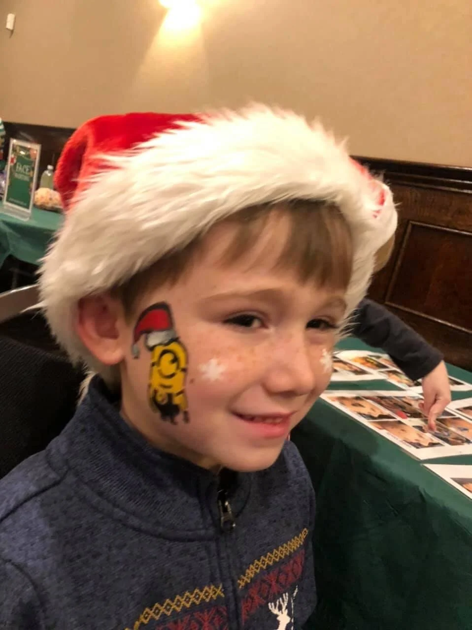 A young boy wearing a Santa hat with face paint of a yellow tiger with a red Santa hat, smiling at a holiday gathering.
