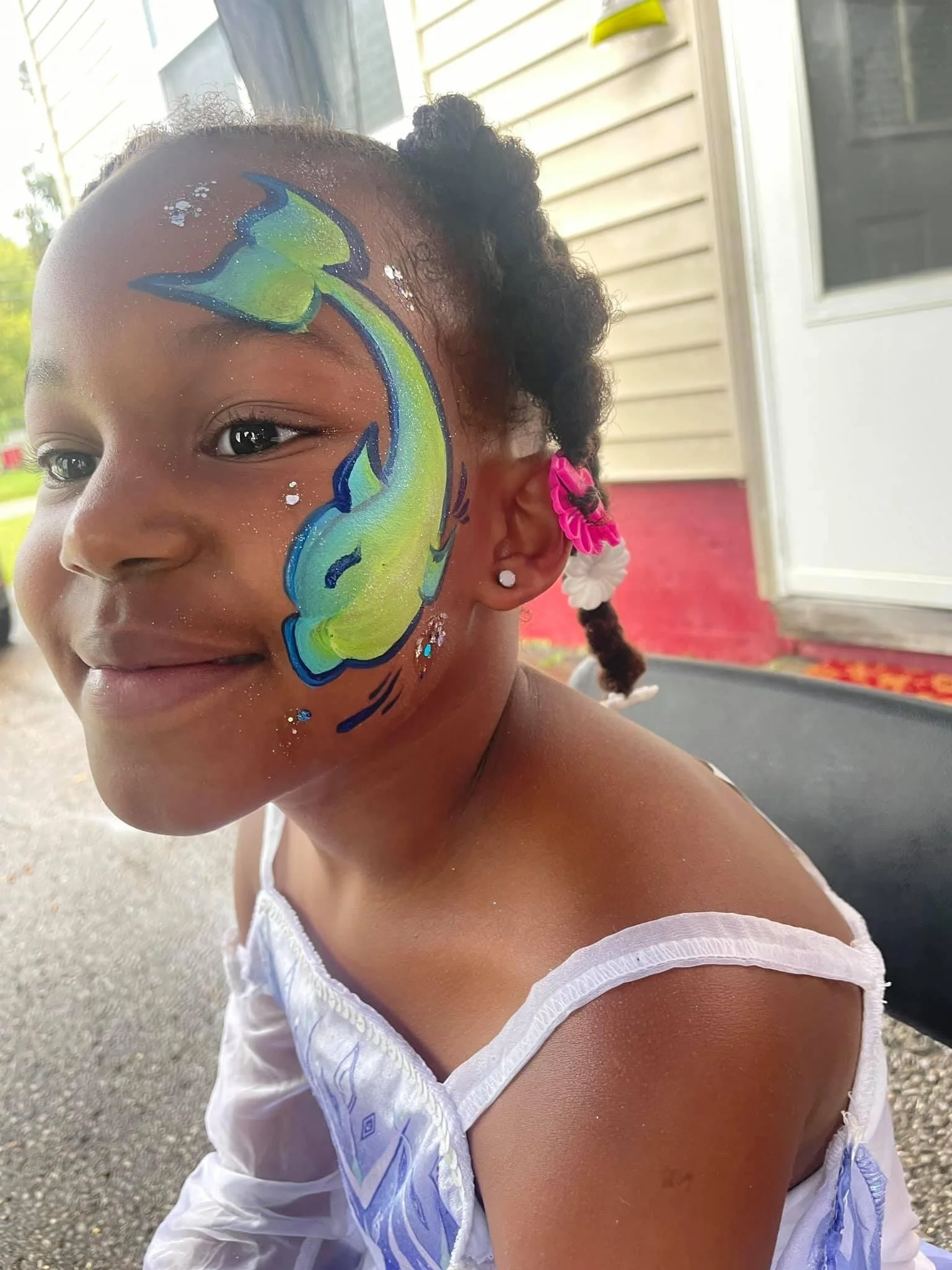Young girl with face paint of a colorful fish on her cheek, wearing a white dress with blue patterns, pink and white hair accessories, sitting on a porch.