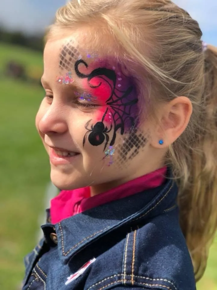A young girl with blonde hair tied back, wearing a denim jacket and a pink hoodie, has face paint of a black spider and web design with pink, purple, and glitter accents on her cheek and forehead, outdoors on a sunny day.