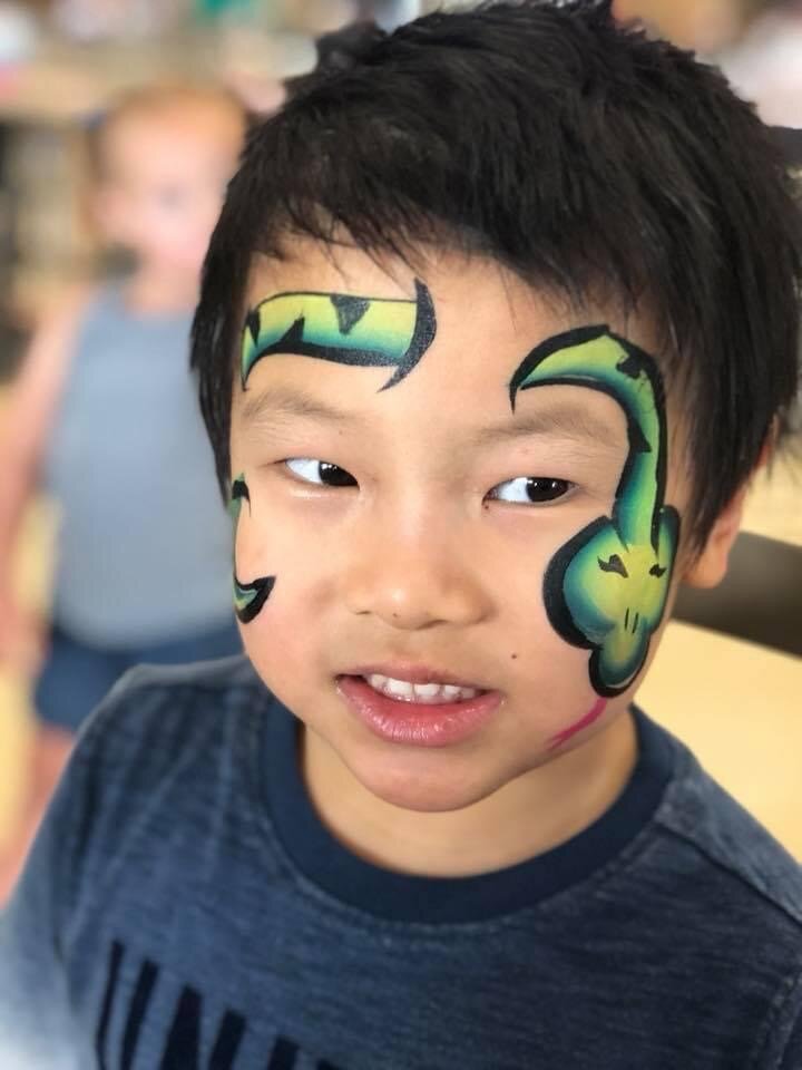 A young boy with black hair has face paint resembling a green and black dinosaur or dragon on the sides of his face.
