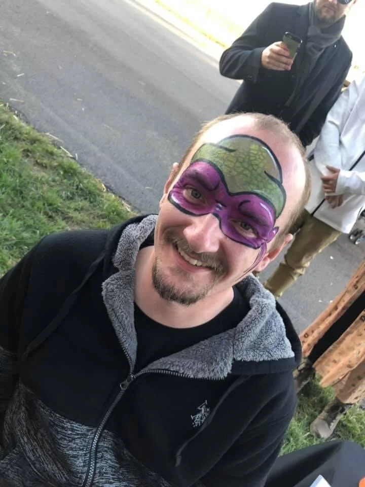 Man with face painted as a purple and green character, smiling outdoors.
