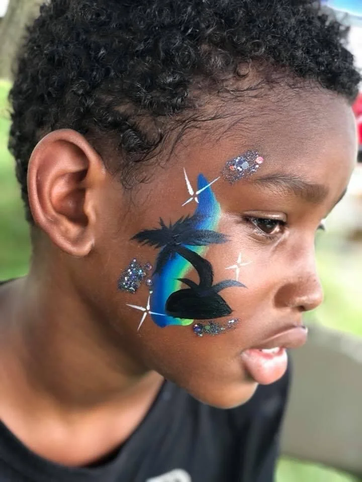 Child with face paint of a black rabbit, palm tree, rainbow, stars, and glitter on his cheek.