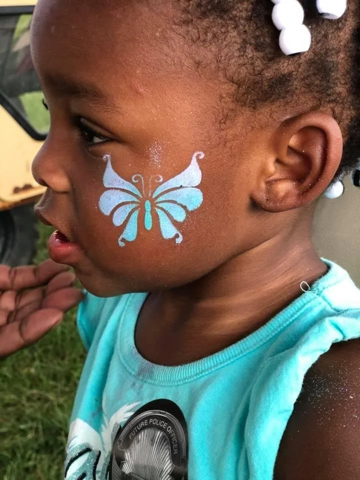 A young African American girl with a painted blue butterfly on her cheek, wearing a turquoise shirt and white beads in her hair, outside during the daytime.