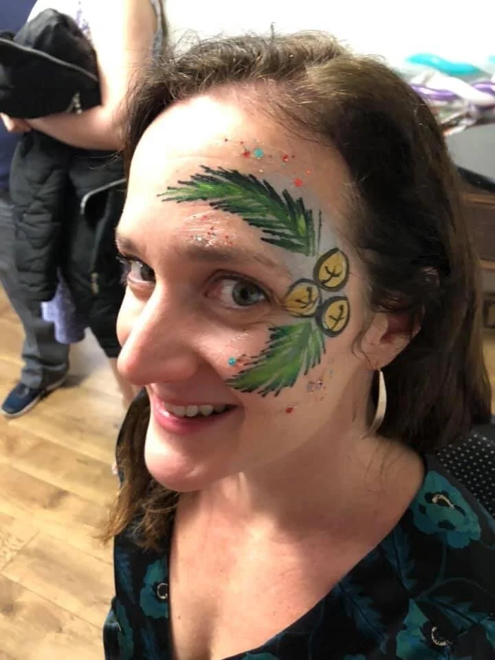 A woman with face paint of a Christmas tree, ornaments, and colorful glitter, smiling indoors.