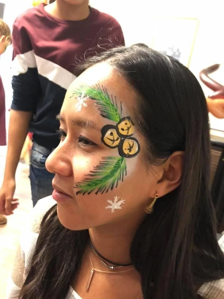A woman with face paint that resembles Christmas ornaments hanging from a holly branch on her cheek, with a white snowflake below. She is smiling with her eyes closed and wearing a black choker and earrings.