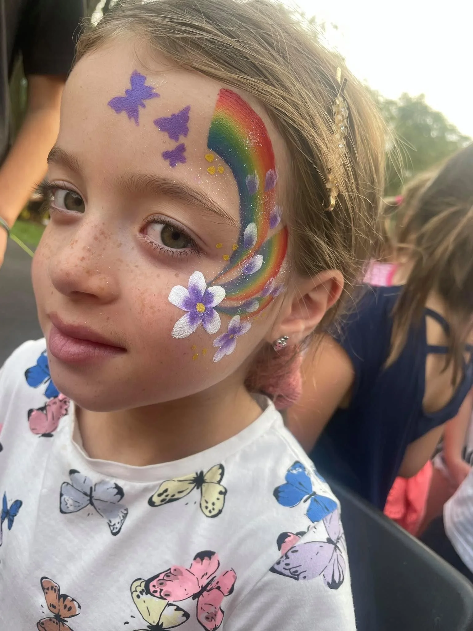 A young girl with face paint of purple butterflies, a rainbow, and purple flowers, wearing a white shirt with butterfly prints.