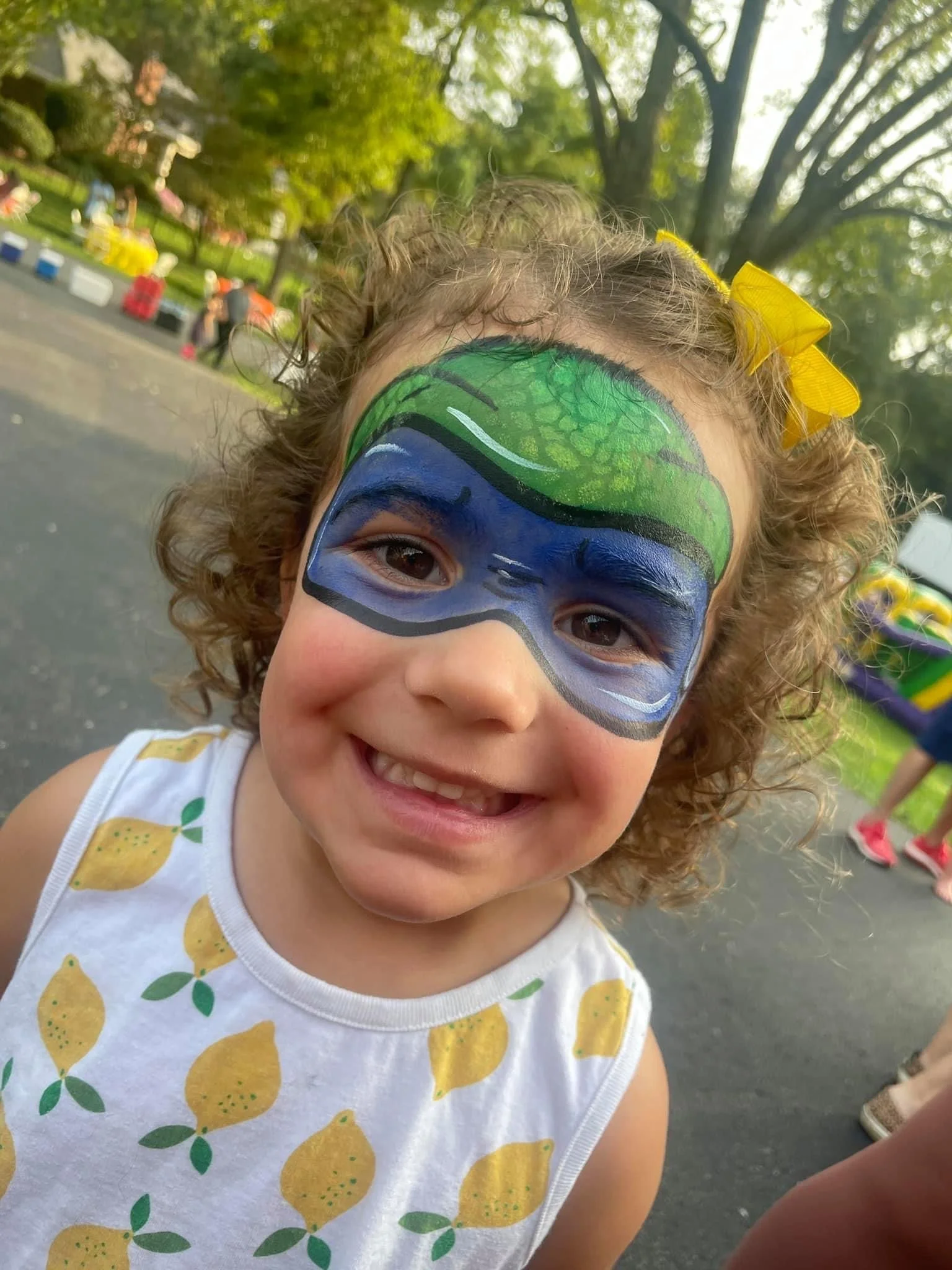 A young girl with curly hair and a yellow bow wearing a face paint design of a fish mask in green and blue with black and white details. She is smiling outdoors, with trees and people in the background.