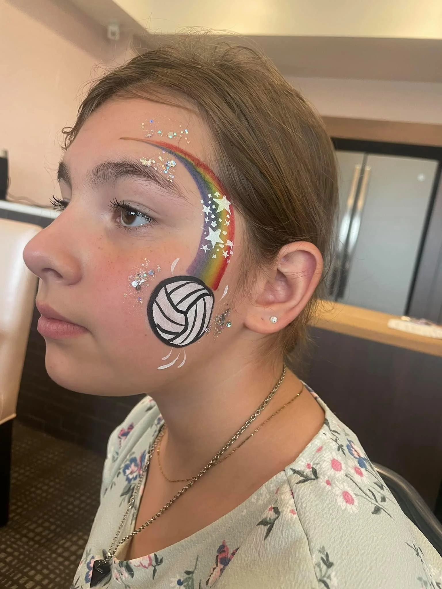 A girl with face paint including a rainbow, stars, glitter, and a volleyball on her cheek, sitting in a room with a refrigerator in the background.