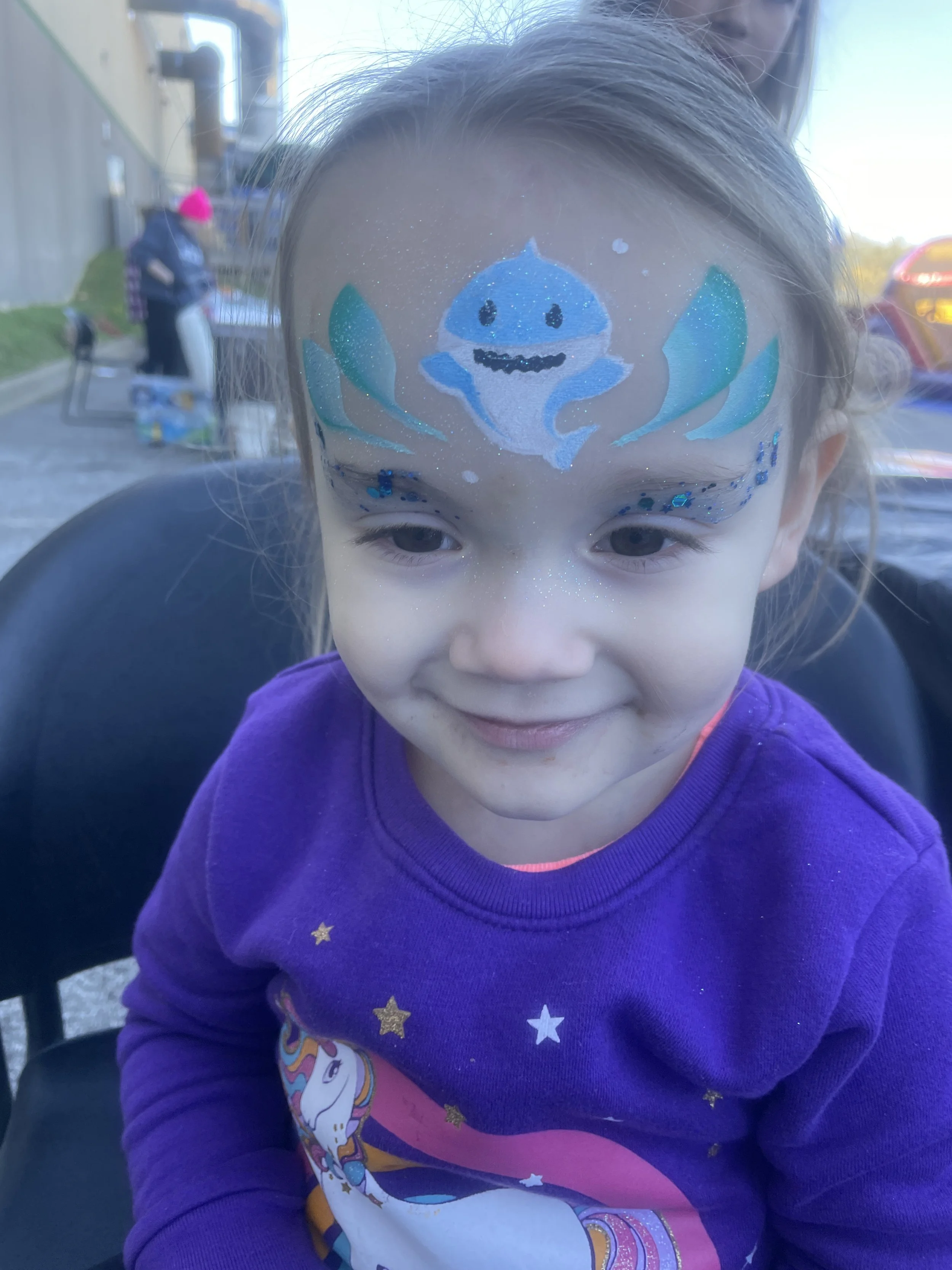 Child with a painted face featuring a blue cartoon crab, glitter, and sea-themed decorations, wearing a purple shirt with a unicorn design.