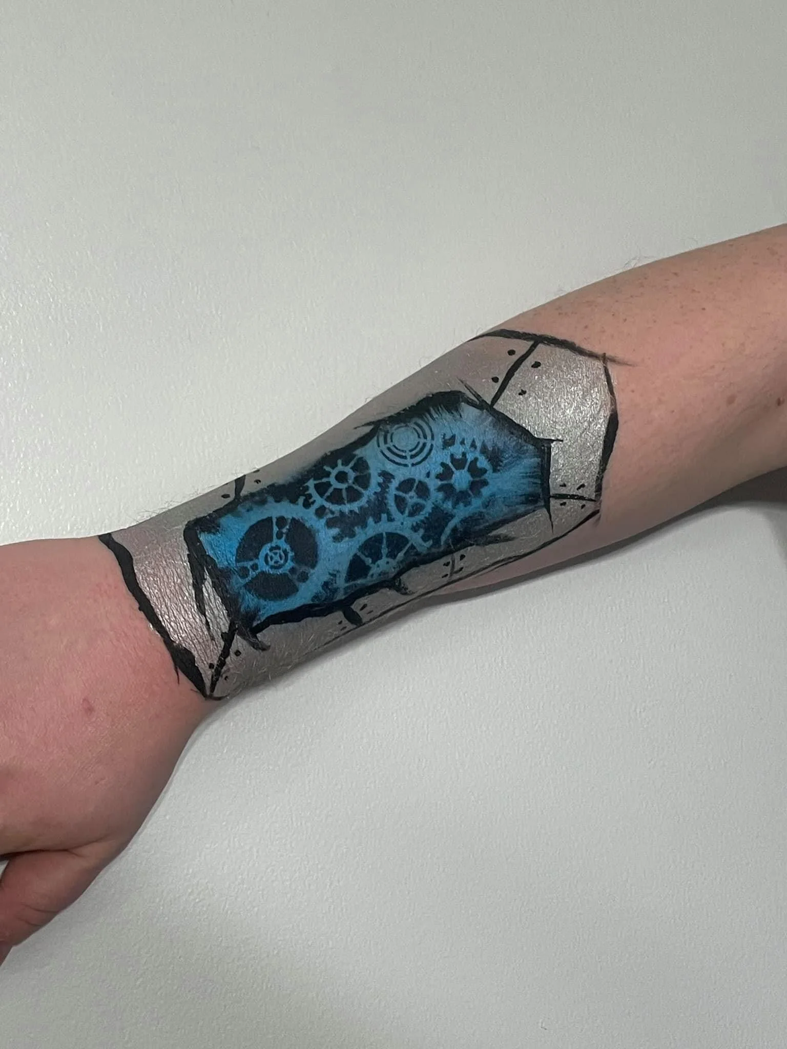 Arm with tattoo of gears and cogs surrounded by a black outline, with parts colored in blue and gray.