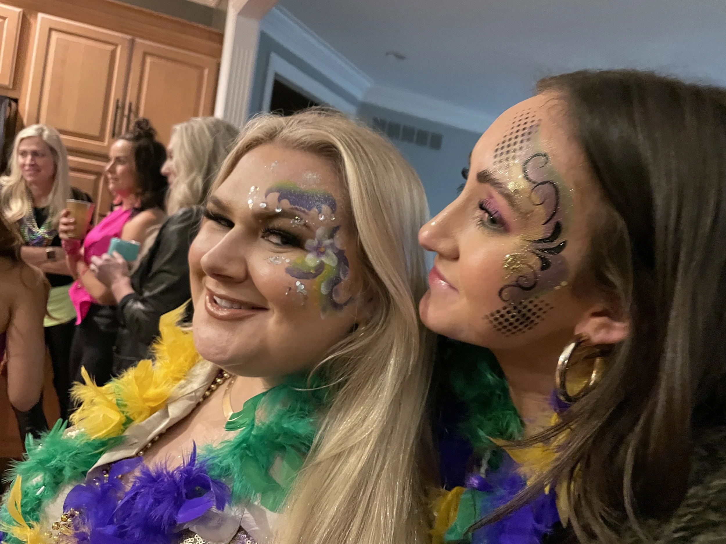 Two women with face paint, colorful feathers, and makeup at a lively party or celebration.