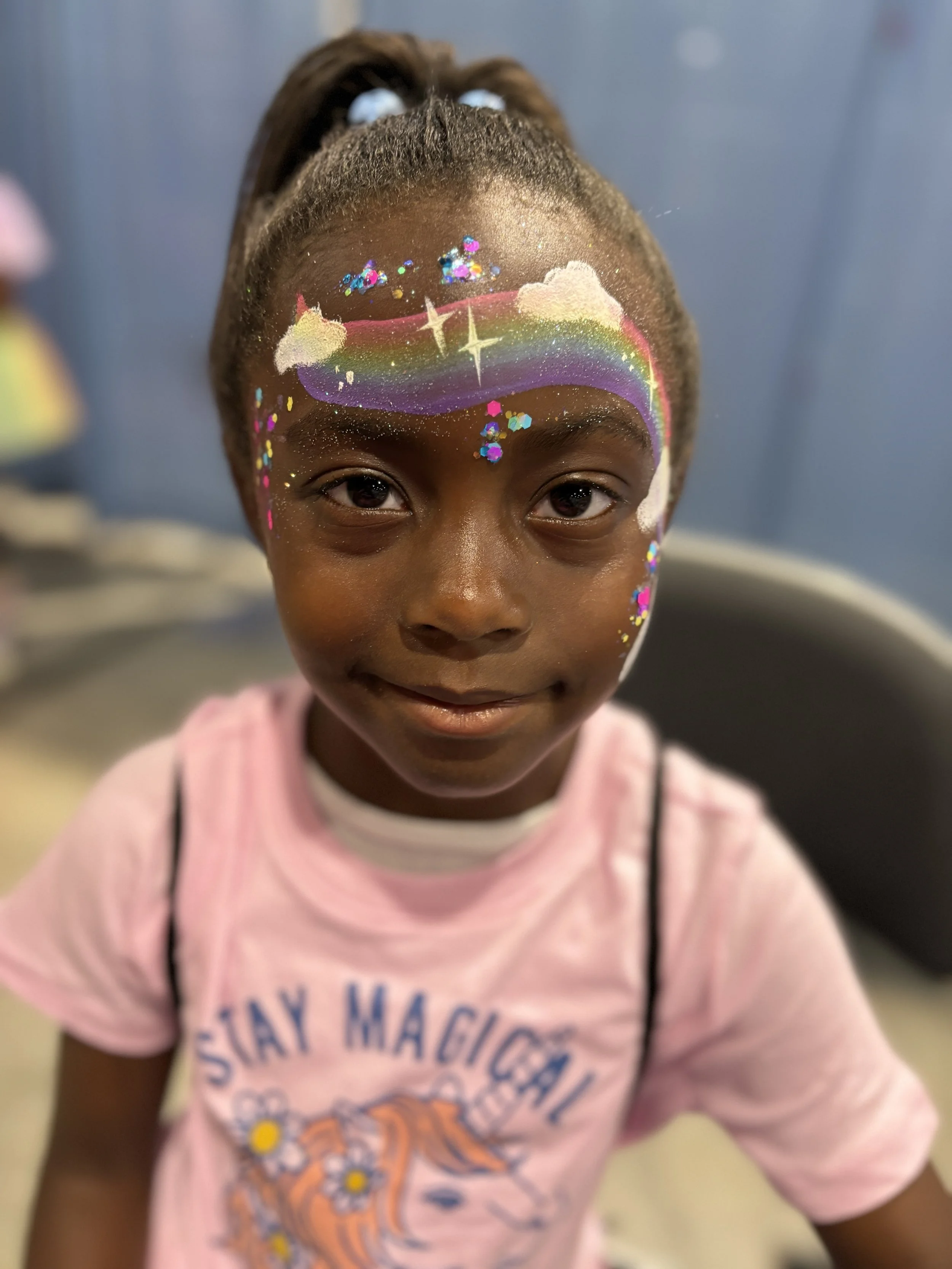 A young girl with dark skin and facial rainbow and cloud face paint, smiling, wearing a light pink t-shirt with a rainbow and the phrase 'Stay Magical'.