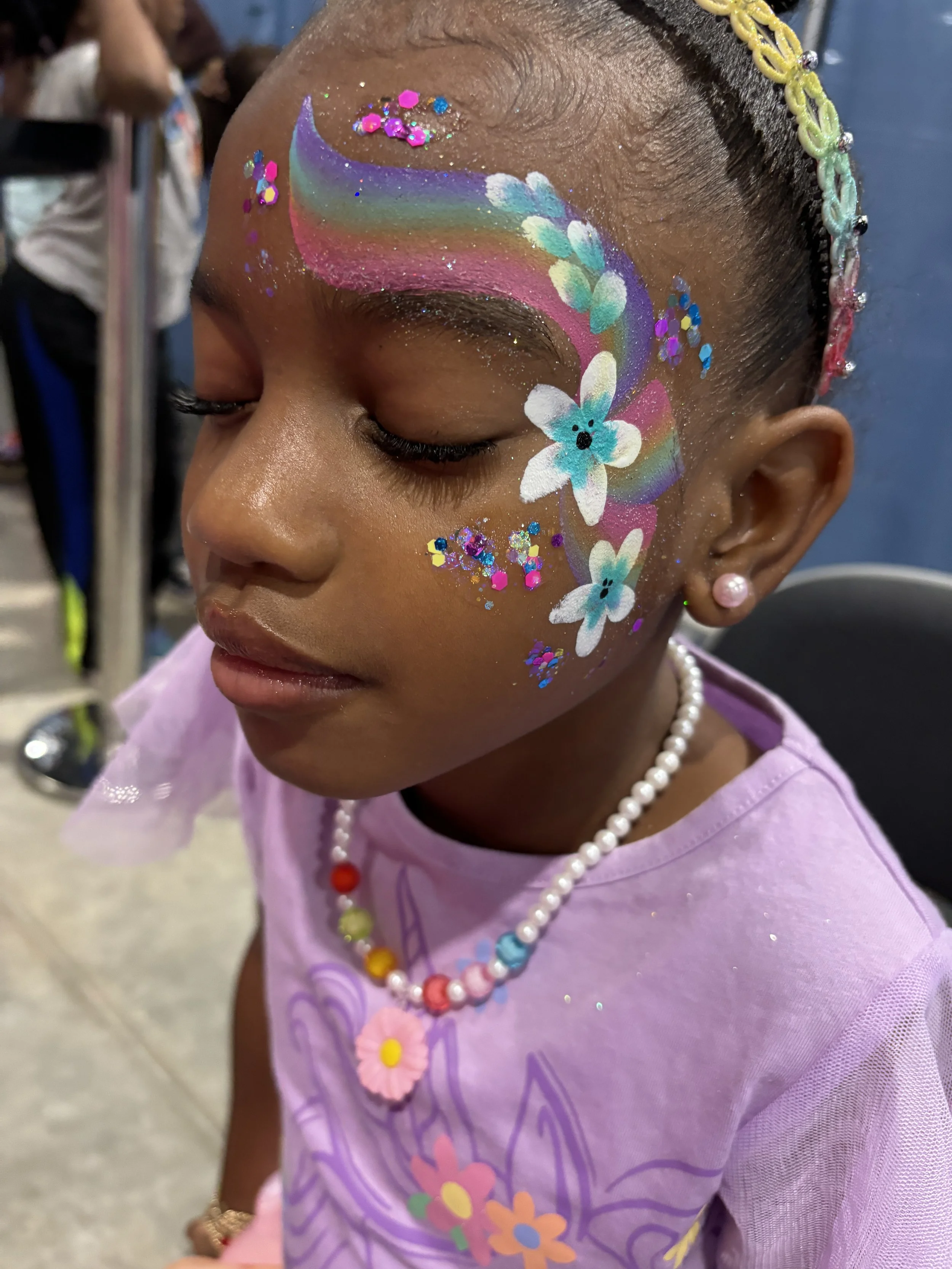 A young girl with face paint featuring a rainbow, white flowers with blue centers, and colorful glitter. She has pearl earrings, a beaded necklace with a flower pendant, and a lavender shirt with a floral design. Her hair is decorated with beads.