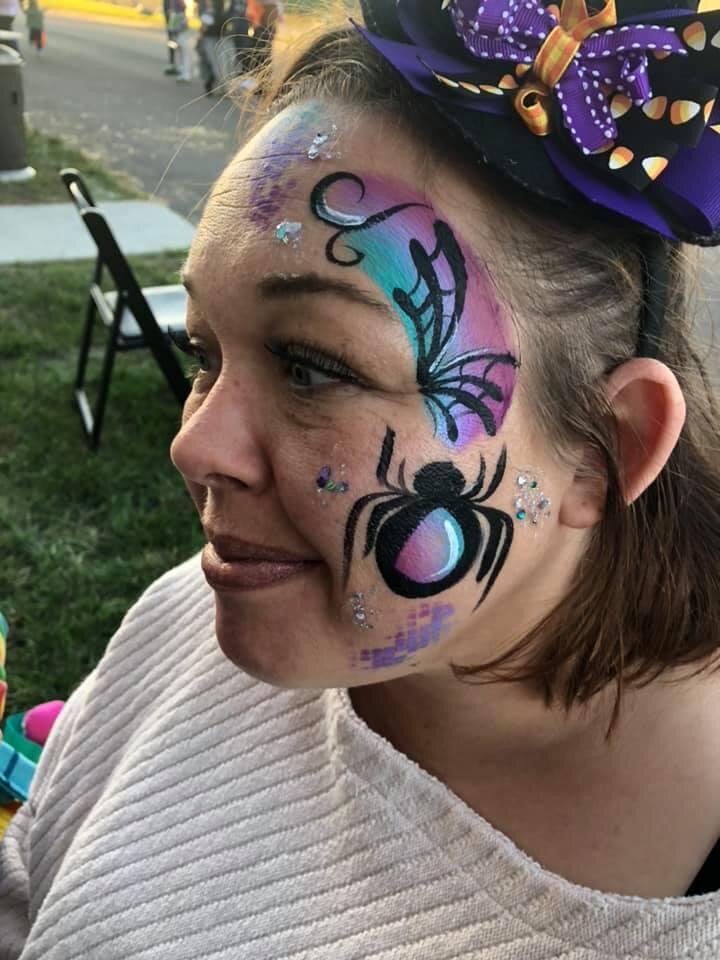 Close-up of a woman with face paint featuring a purple and blue butterfly, a black spider, and rhinestones, wearing a purple polka-dot bow in her hair, outdoors at an event.