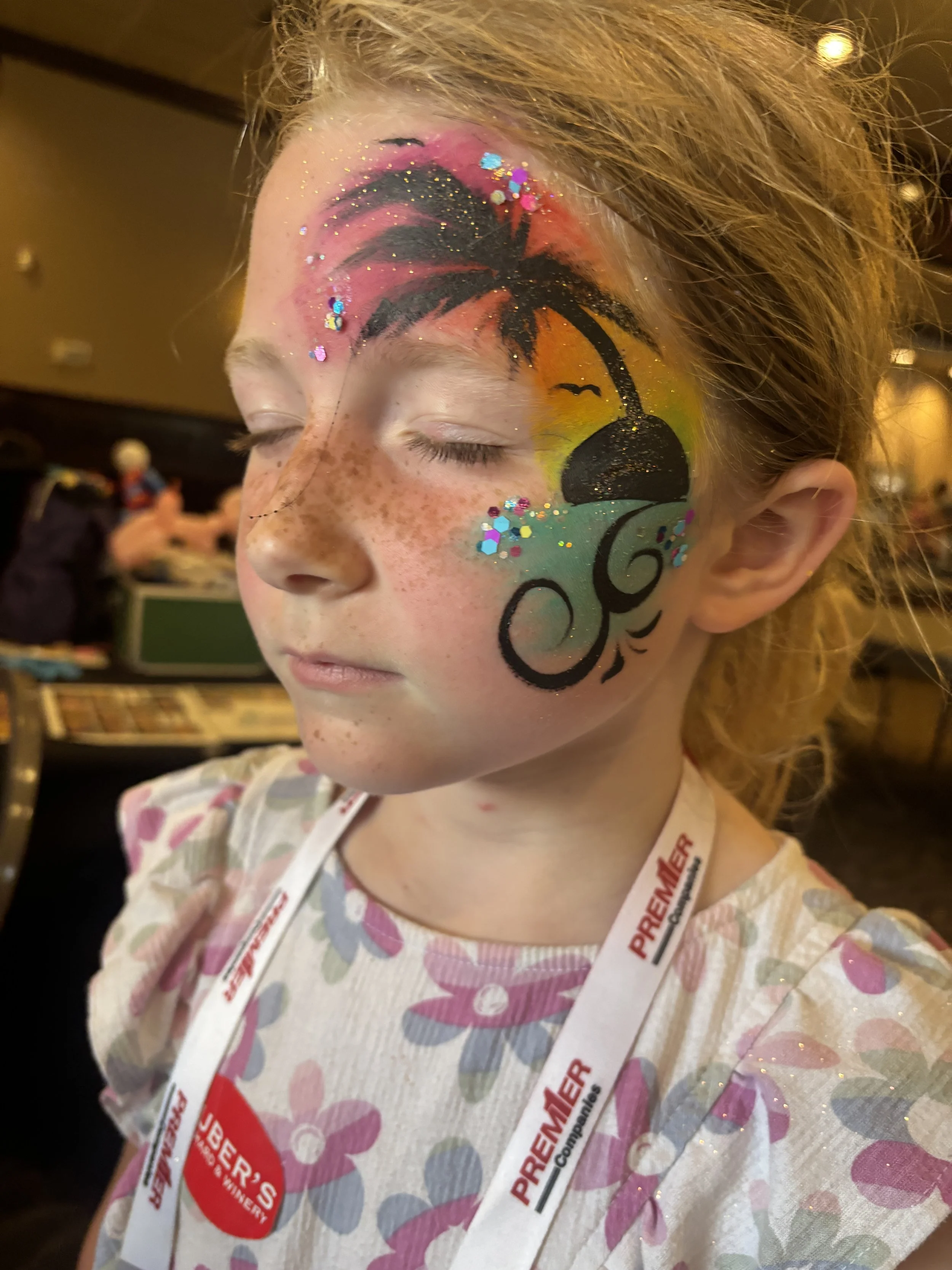 A young girl with face paint of a tropical sunset scene, including a black palm tree, a setting sun, and swirling decorative patterns, with glitter accents, in an indoor setting.