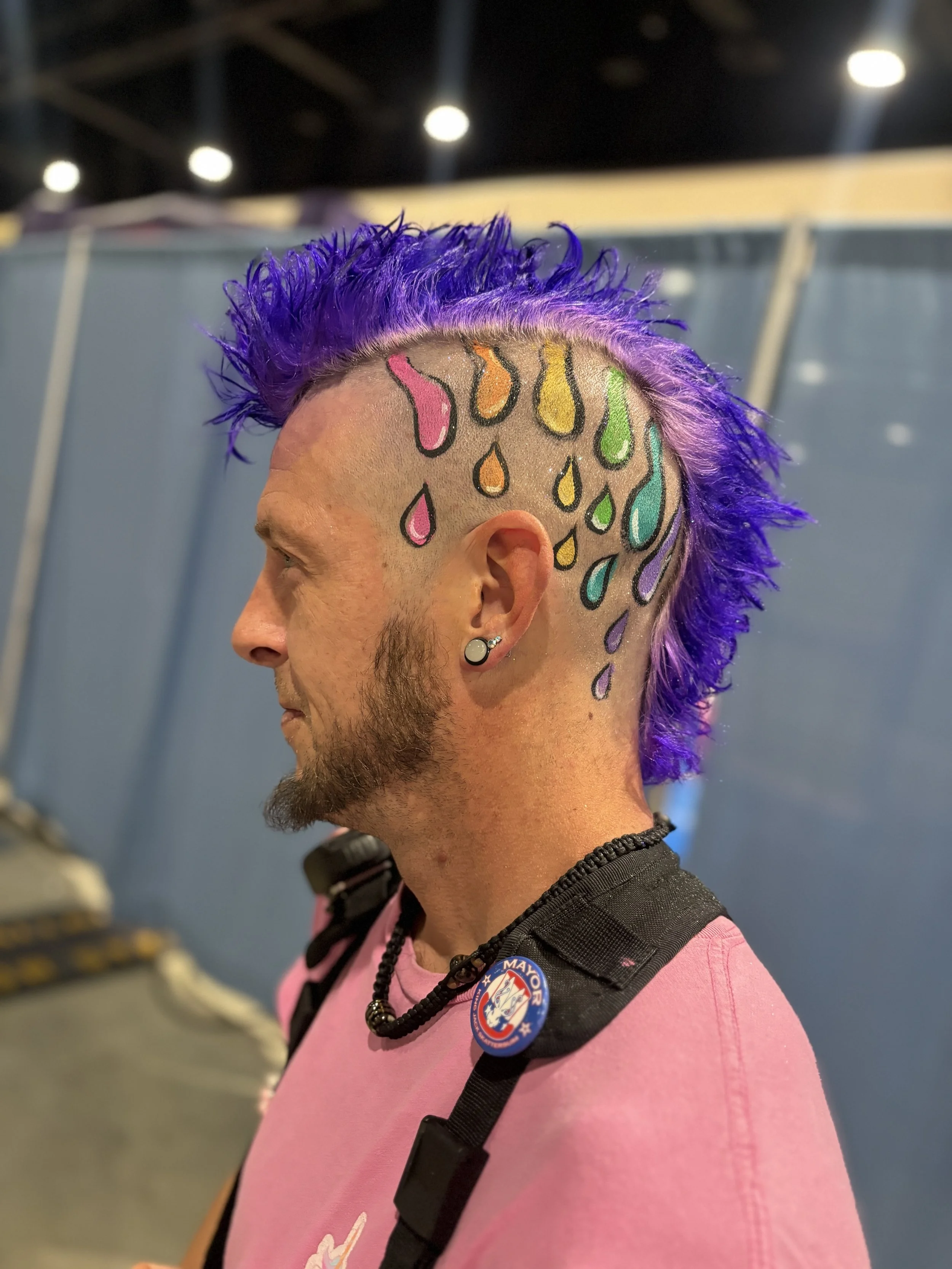 A man with a Mohawk hairstyle dyed purple, with rainbow-colored teardrop tattoos drawn on his head. He has a beard, a body piercing, and wears a pink shirt with black straps. There is a button that says "Mayor" attached to his strap.