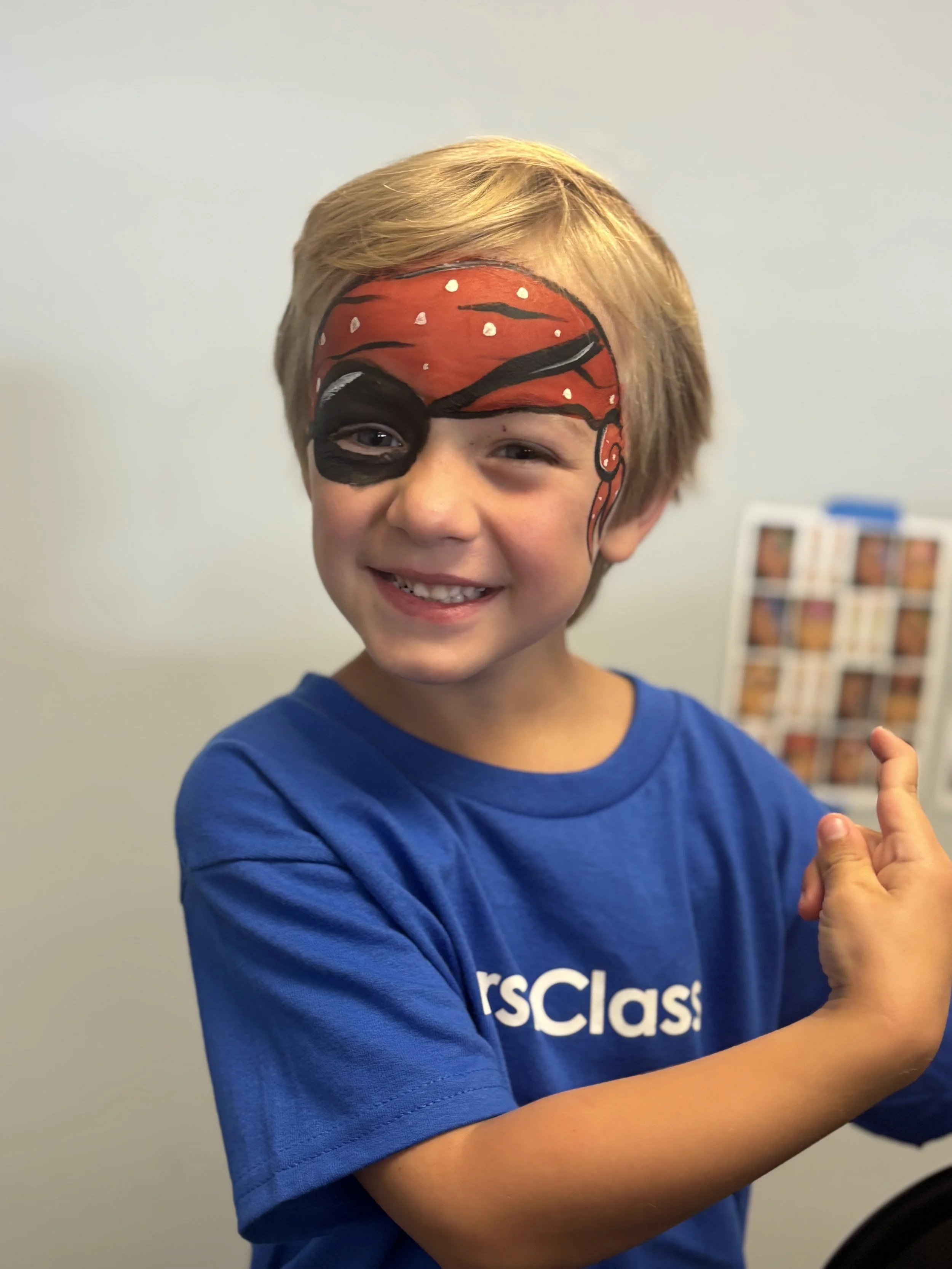 A young boy with face paint resembling a pirate's mask, smiling and pointing to his right, wearing a blue t-shirt.