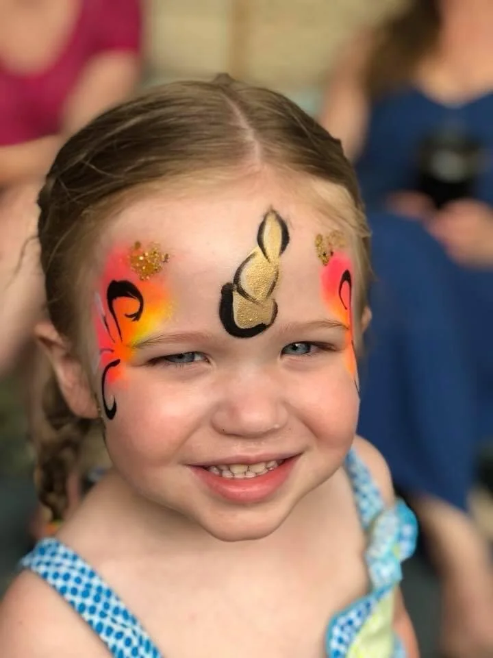 Close-up of a young girl with face paint resembling a butterfly, with black, gold, and red colors, smiling outdoors.