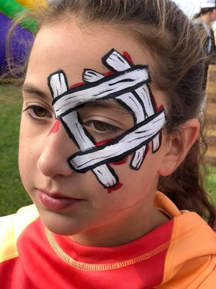 A young girl with face paint depicting a bandaged eye, with black and red details, and her natural eye visible through the painted bandage.