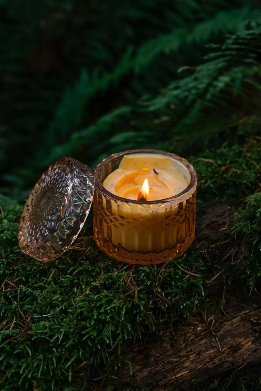 A lit candle in a decorative amber glass container with a matching lid, placed on mossy ground amidst greenery.
