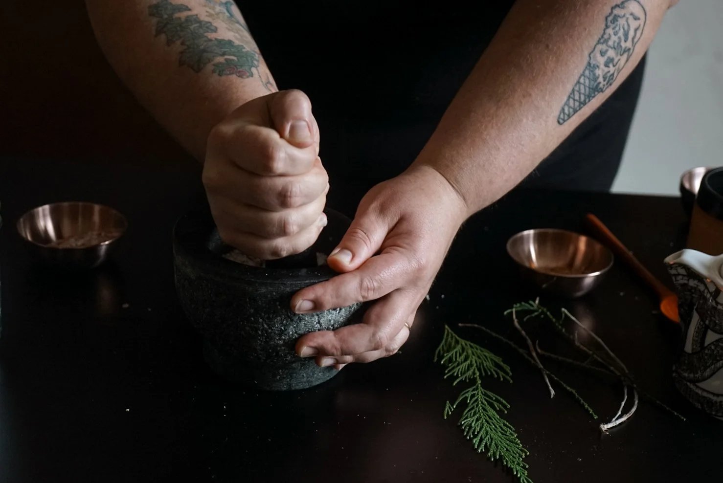 Person using a mortar and pestle to grind herbs on a black surface, with small copper bowls, sprigs of greenery, and a ceramic mug nearby.