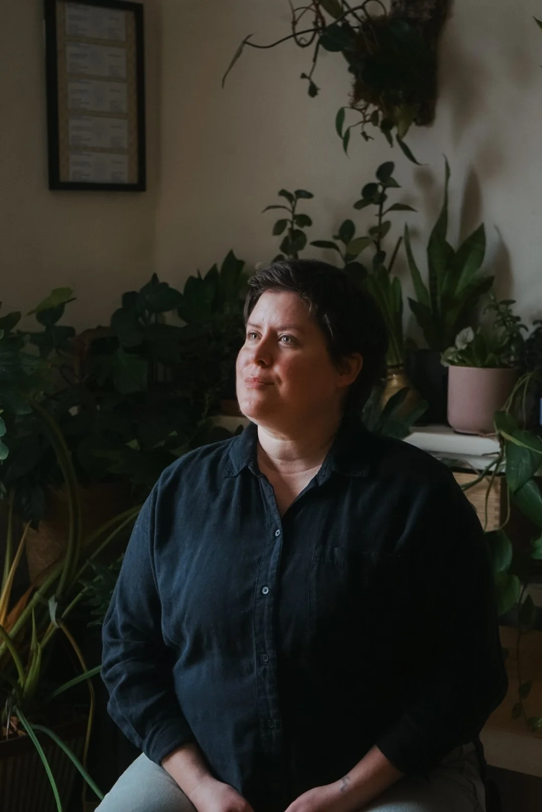 A woman with short dark hair, wearing a black shirt, sits in a room filled with houseplants and looks thoughtfully to the side.