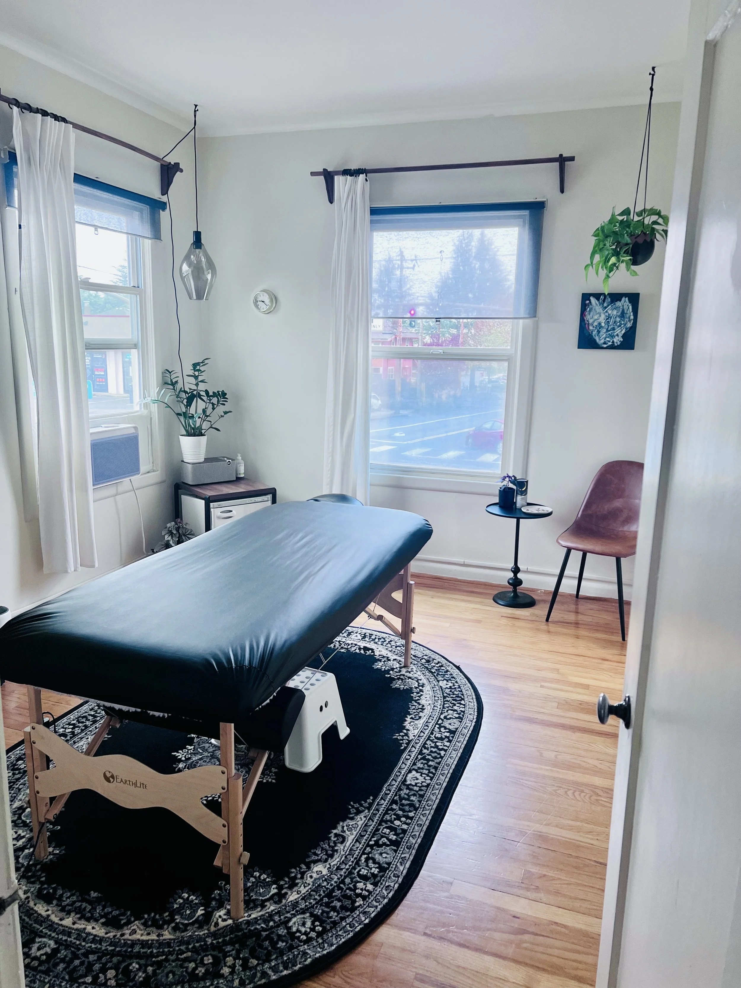 A massage therapy room with a massage table, black cover, and wooden legs, situated in front of a window with sheer and blackout curtains. The room has hardwood floors, a black patterned rug, a small side table with candles and a plant, a pink chair,