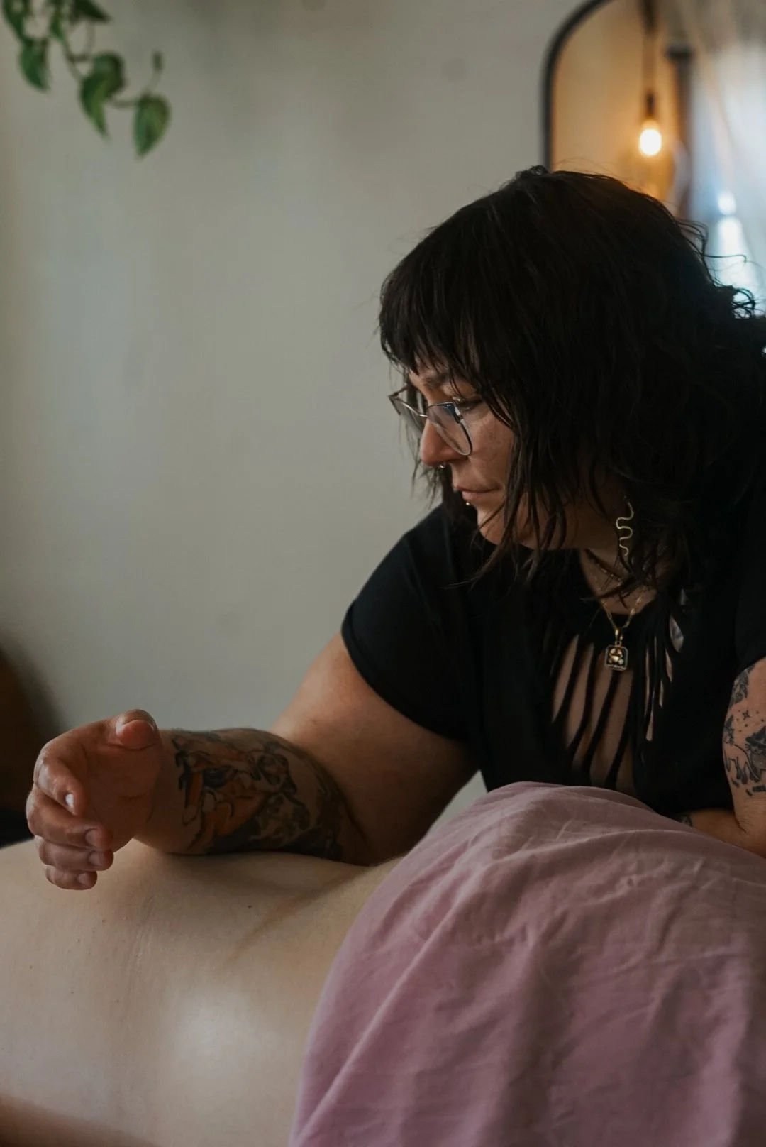 A woman with dark hair, glasses, and tattoos on her arms gives a massage to a person lying on a bed.
