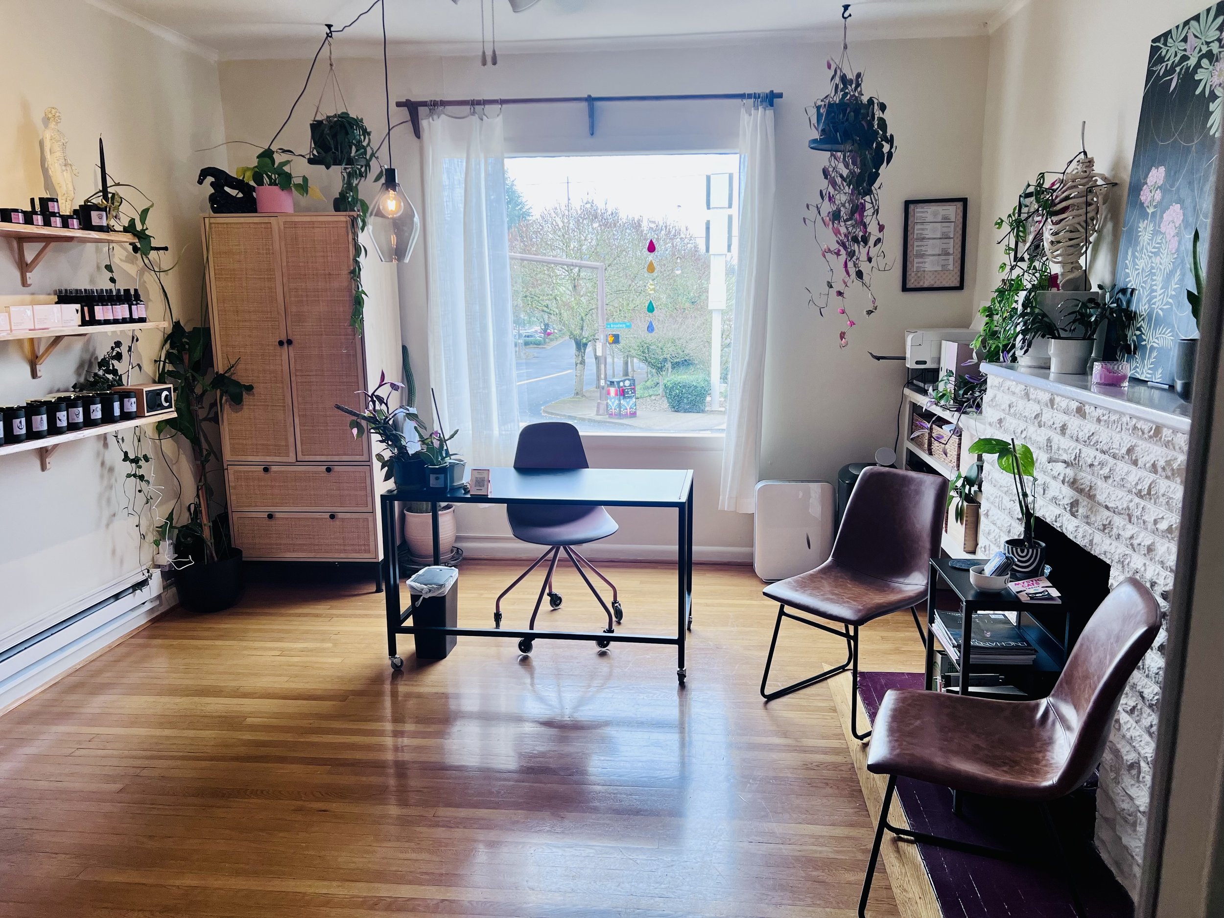 A cozy, well-lit room with wooden floors, a large window with white curtains, and several plants. There is a desk with a chair near the window, a shelf with plants on top, and a seating area with two chairs in front of a white brick fireplace.