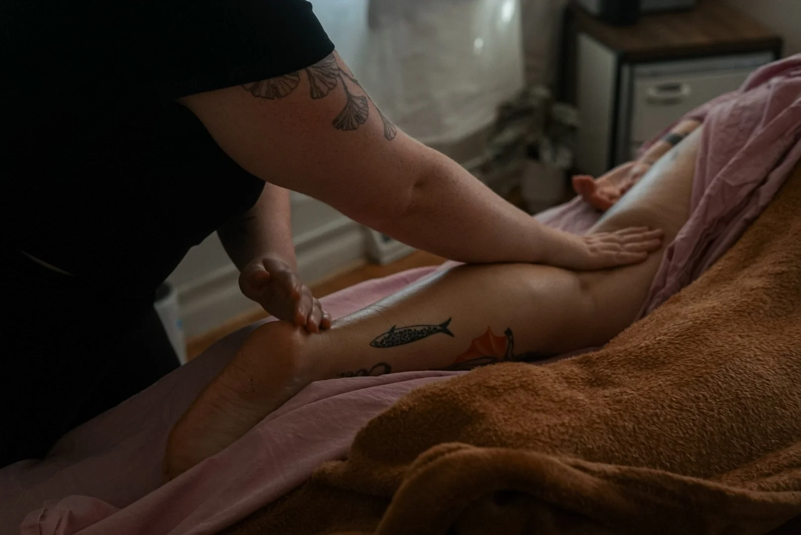 A person receives a massage on their leg from a massage therapist in a dimly lit room, with a bed covered in pink and brown towels, and a small wooden nightstand in the background.
