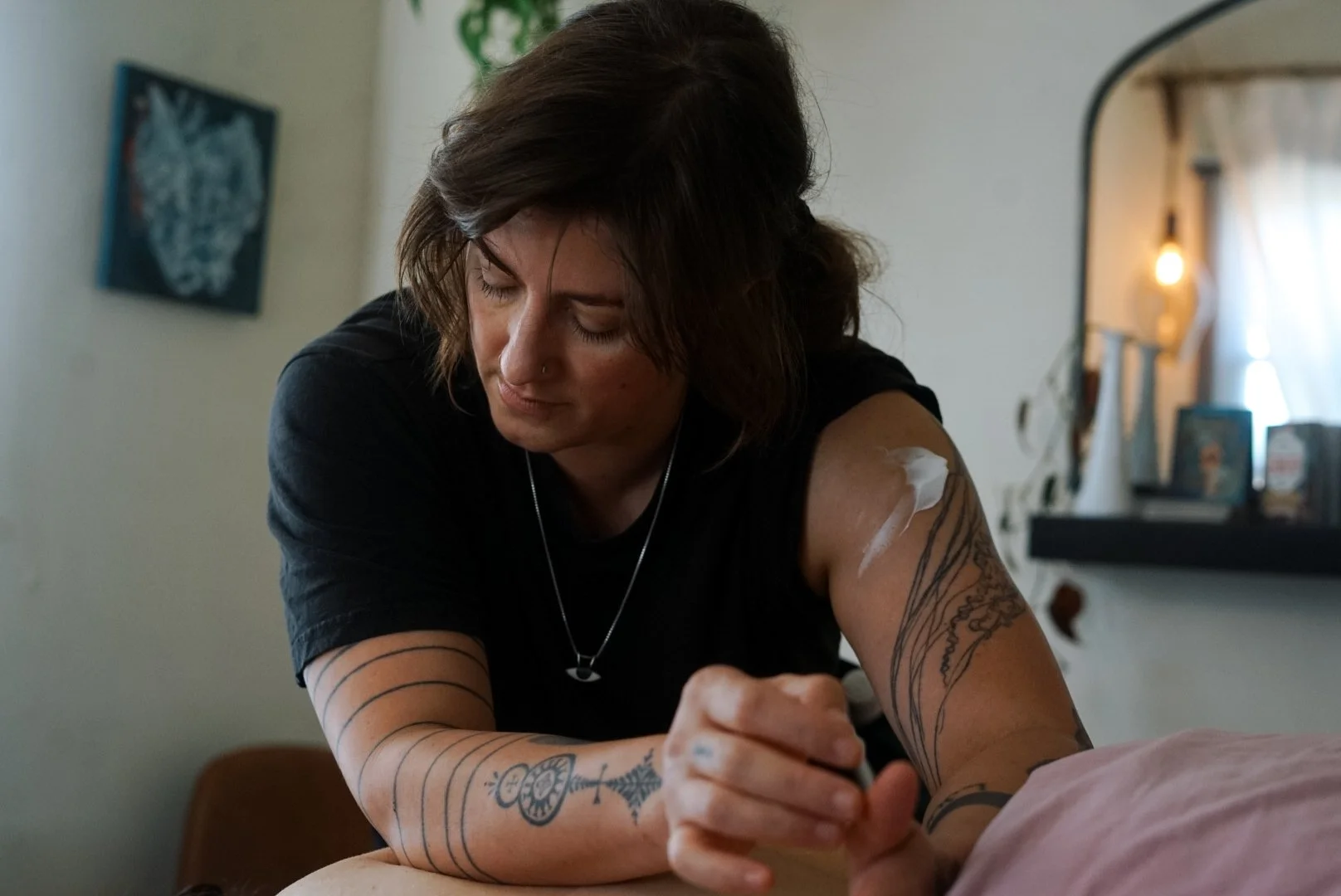 A person with tattoos on their arms and a nose piercing is applying lotion or cream to their shoulder. They have shoulder-length brown hair and are wearing a black shirt and a silver necklace.