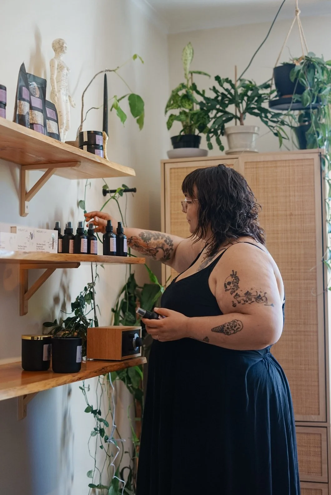 A woman with tattoos and glasses wearing a black dress, selecting essential oils or bottles from wooden shelves in a room with potted plants.