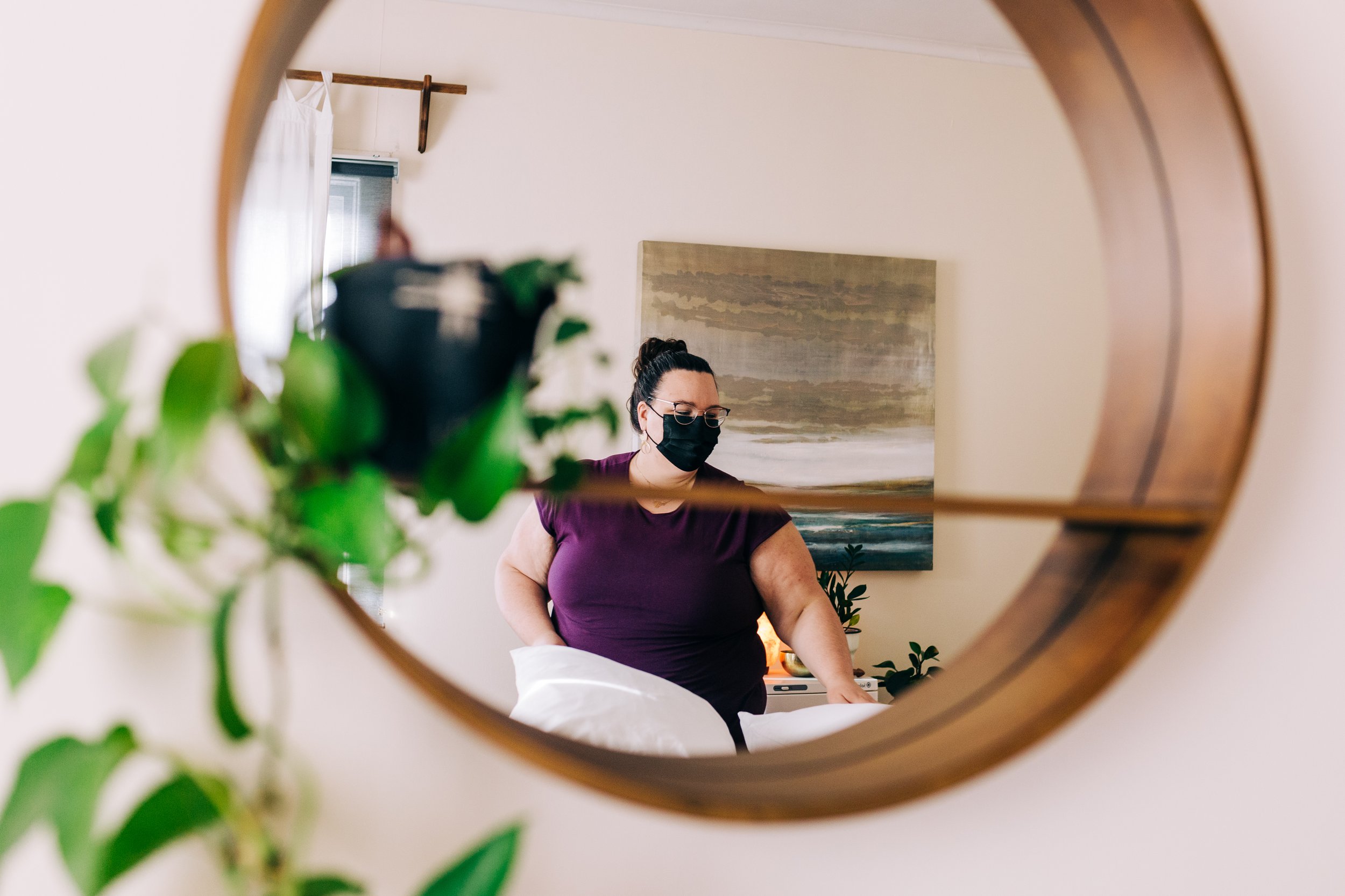 Person wearing a mask and glasses, reflected in a round mirror with plants and artwork in the background.