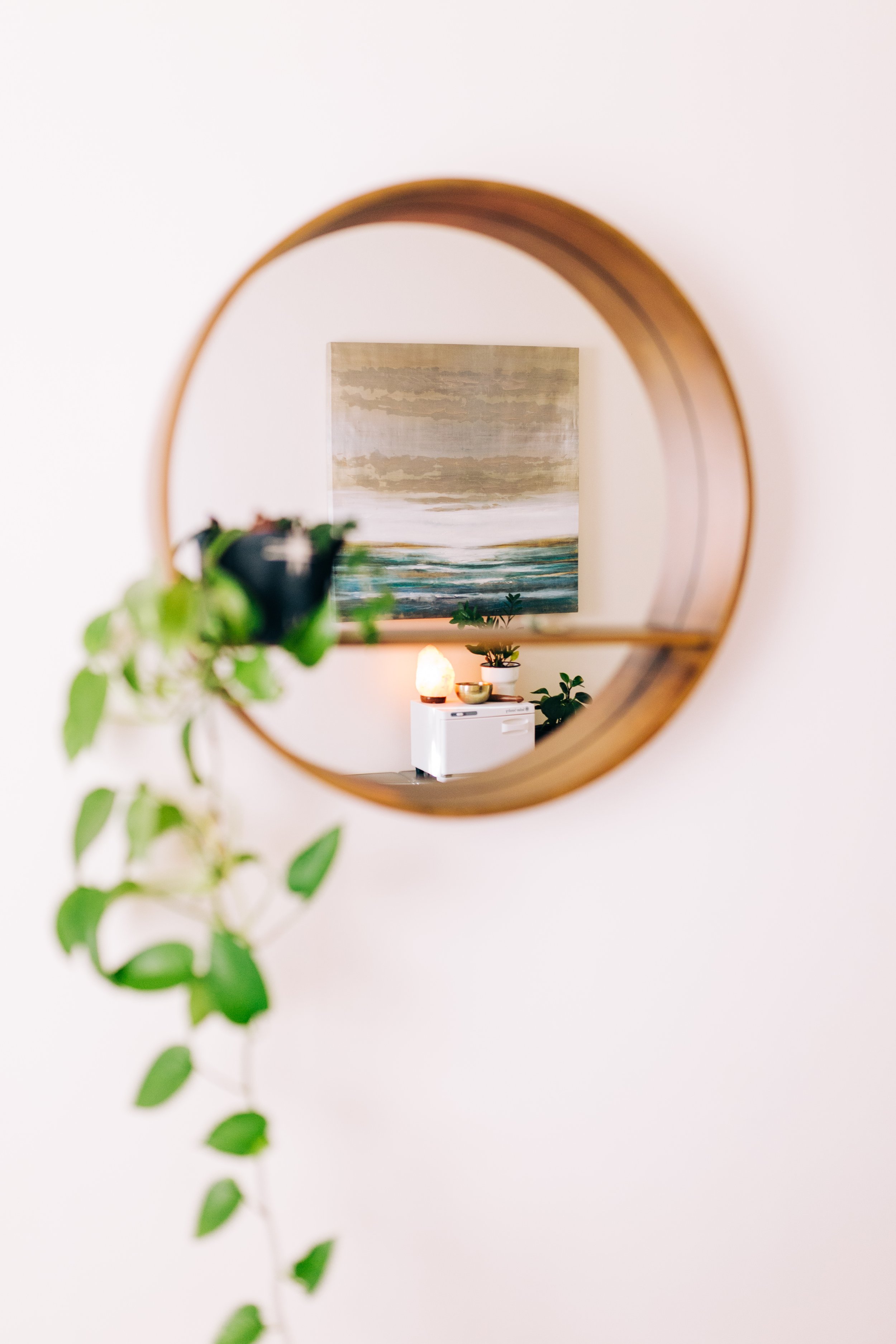 Round wall mirror reflecting a painting, a Himalayan salt lamp, potted plants, and a small white appliance; vine plant on the left.