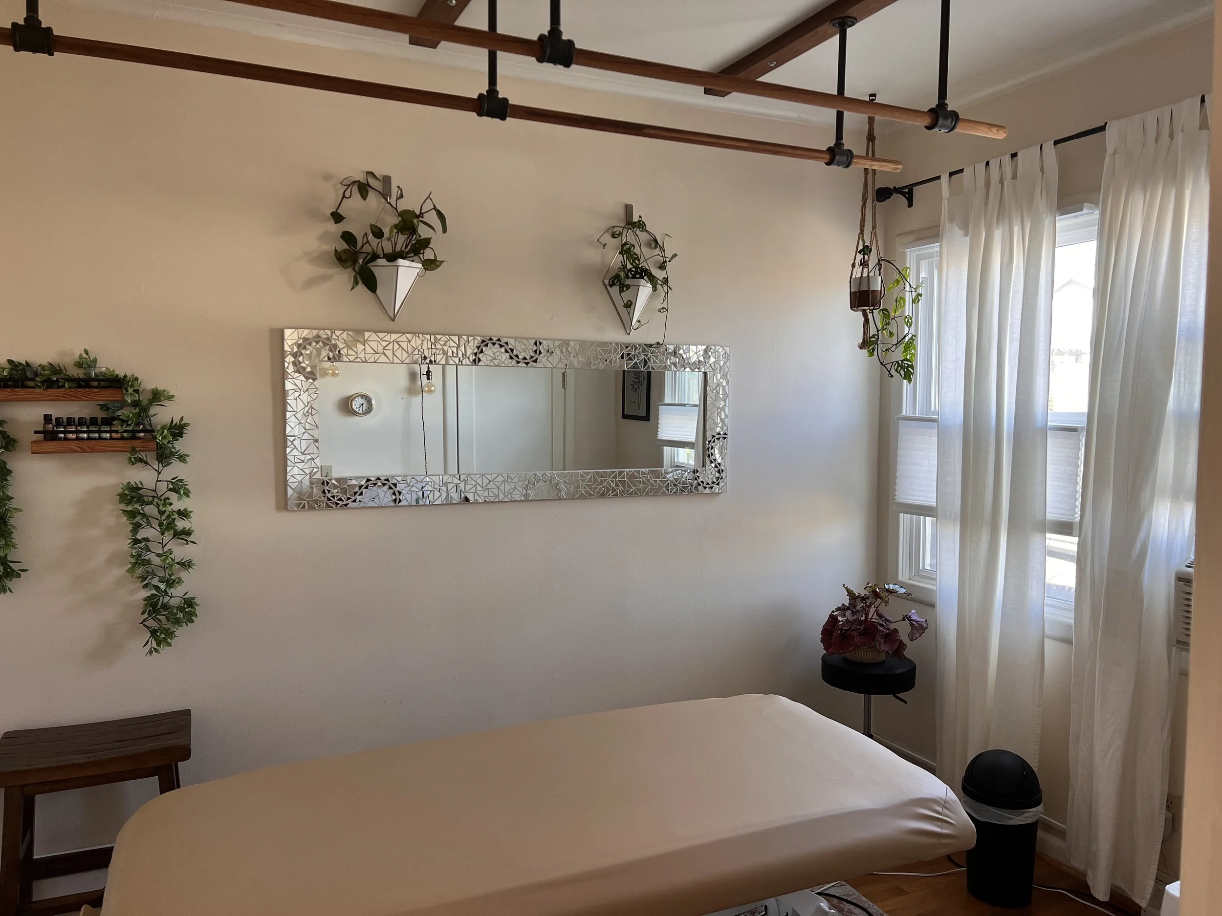 A cozy room with white walls, decorated with hanging plants, a large mirror, and a massage table covered with a cream sheet. There are wooden ceiling beams, white curtains over the windows, and a small side table with flowers and a black waste bin.