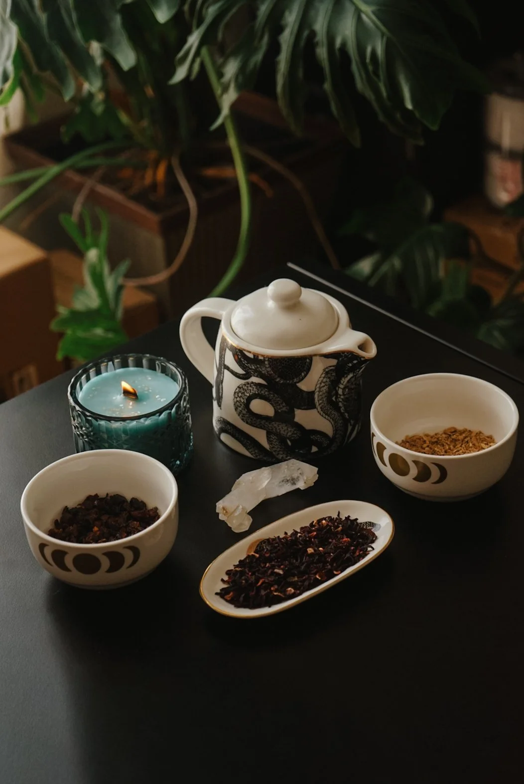 A table with a black surface holding a teapot decorated with black snakes, two bowls of dried herbs or tea leaves, a blue candle, a piece of white crystal, and a small oval dish with dark loose tea leaves. In the background, there are green houseplan