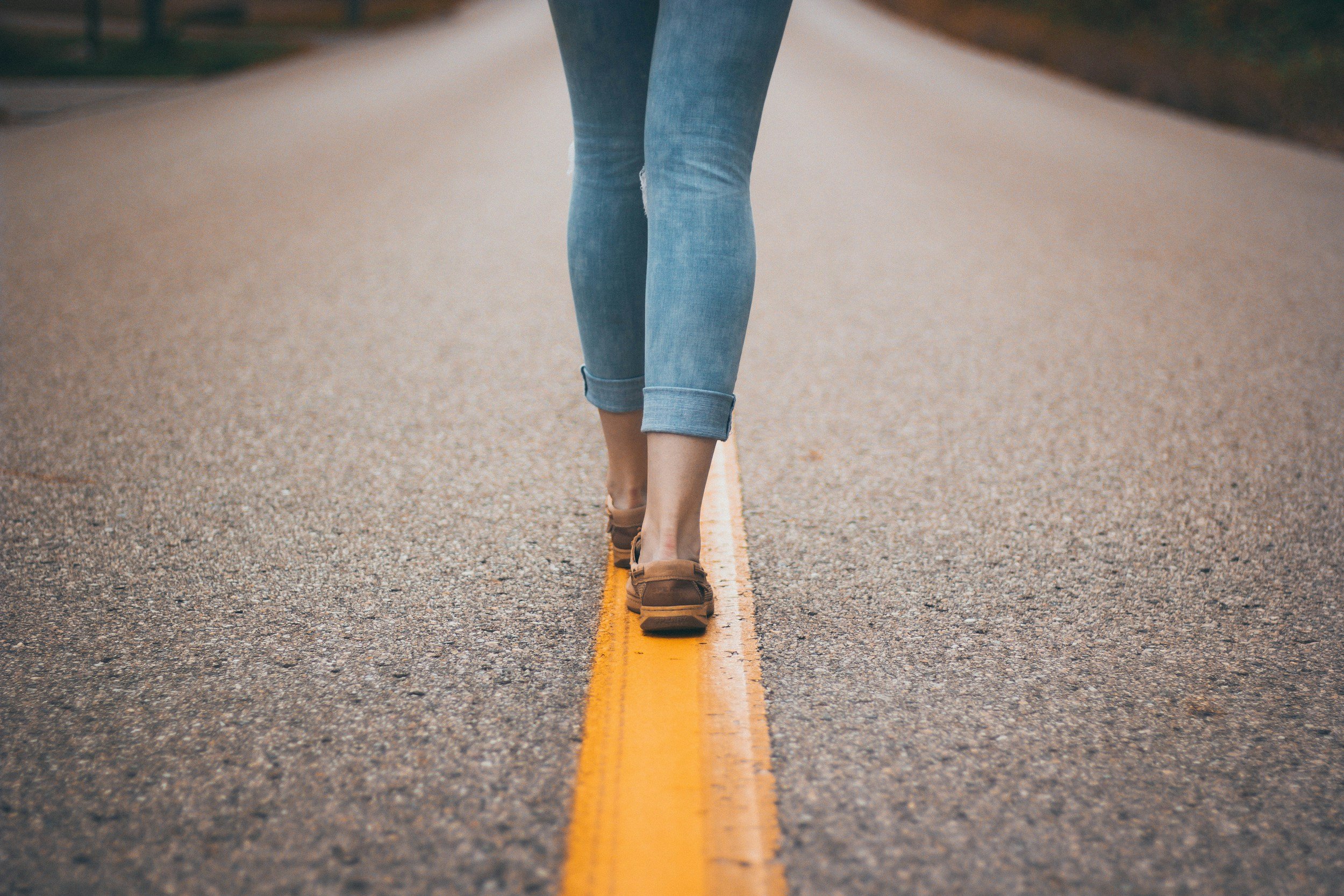 Person walking on a road with yellow lines, wearing rolled-up jeans and brown shoes.