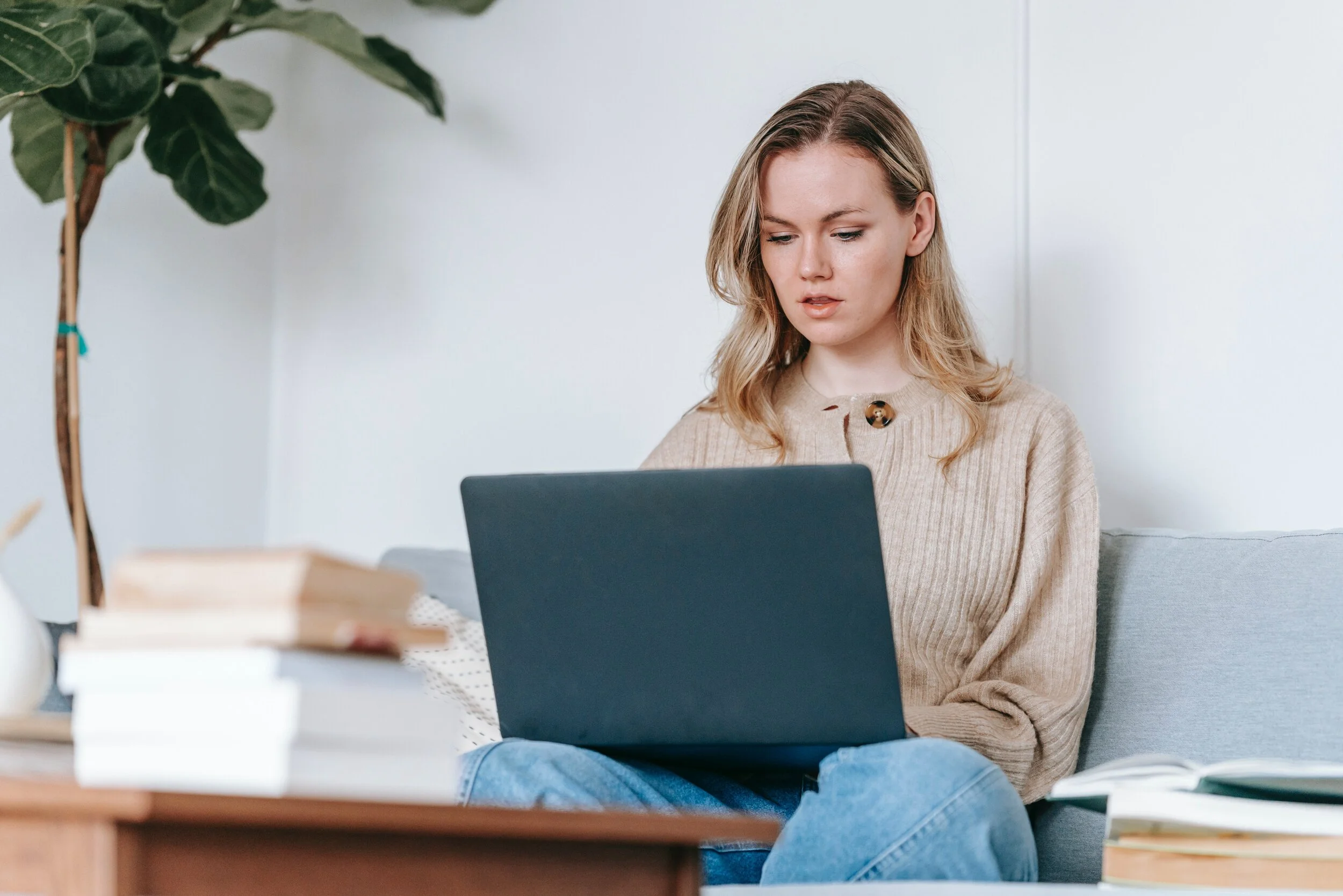 Woman sitting on a sofa working on a laptop with books on a table in front of her.