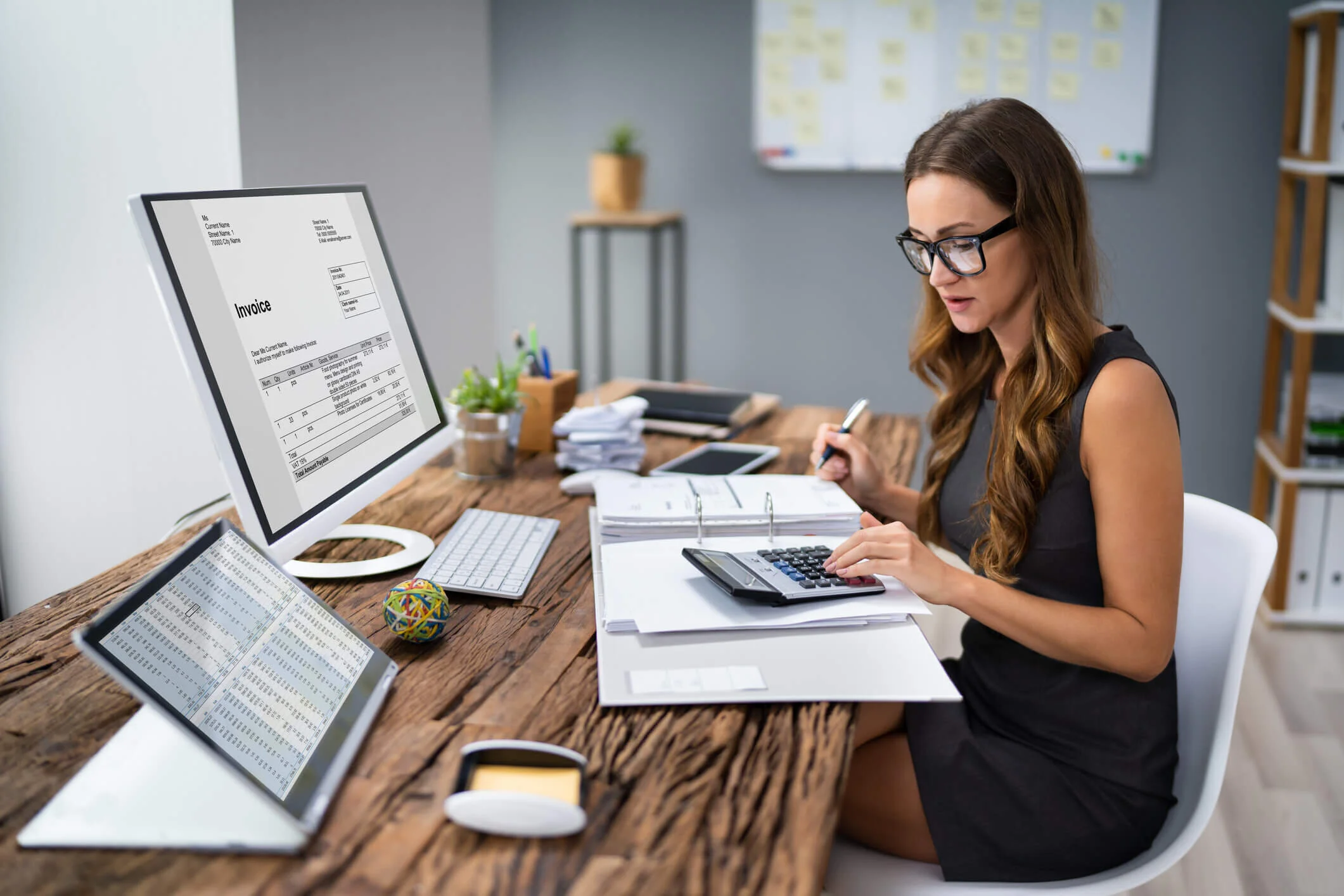 Woman in glasses working on financial documents at a wooden desk with a computer, calculator, tablet, and office supplies in a modern office.