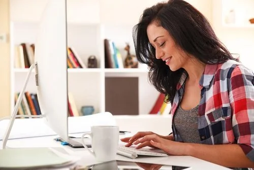 A young woman with dark hair working at a desk with a computer and a coffee mug in a tidy room with bookshelves.